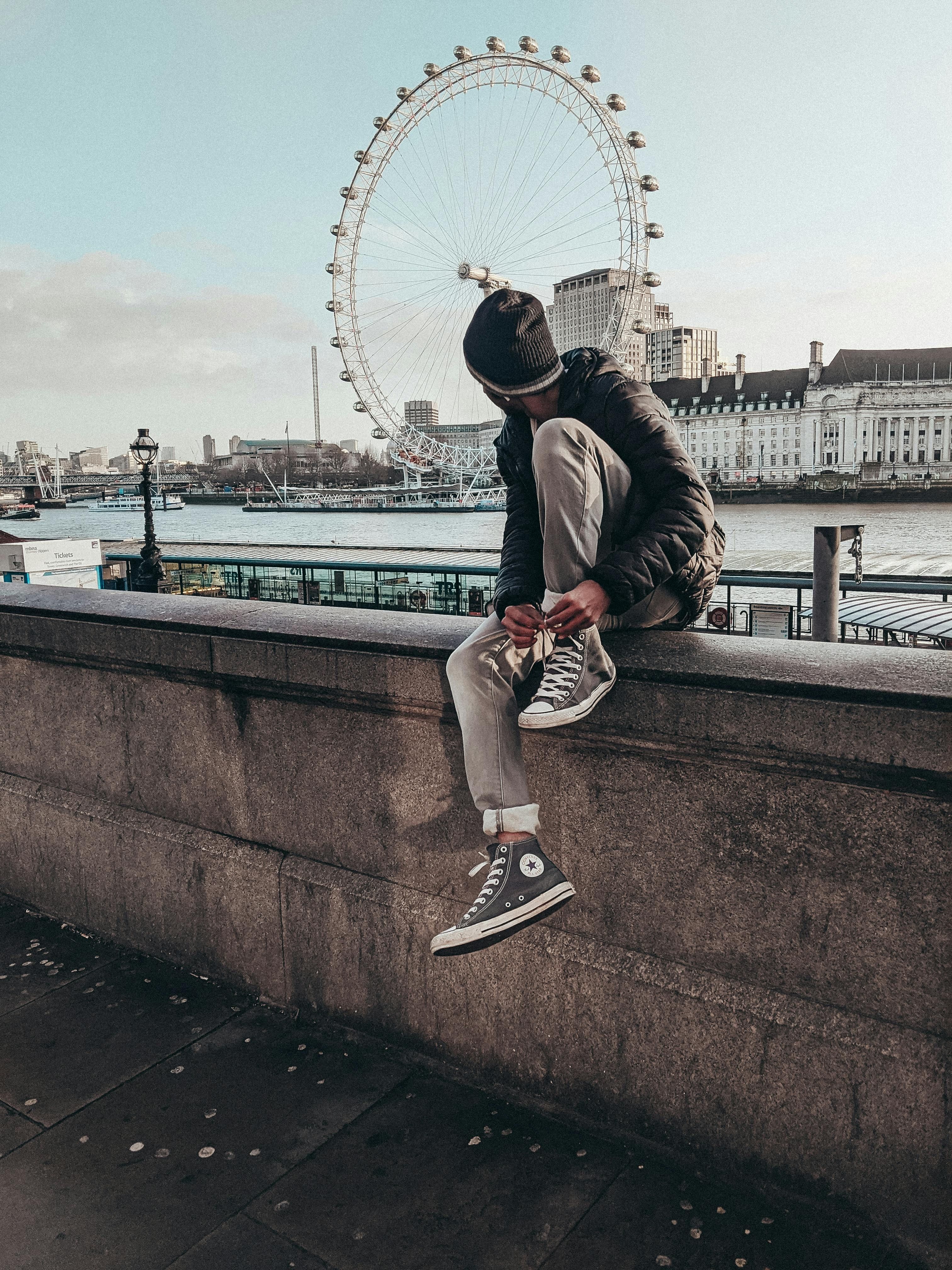 En ung mann sitter på en mur og ser på London Eye i London.