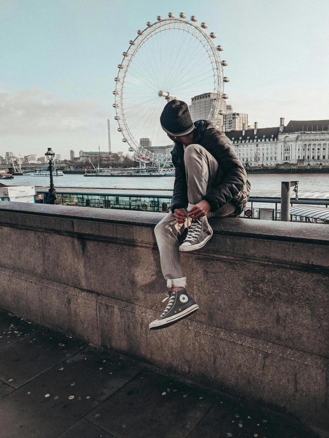 En ung mann sitter på en mur og ser på London Eye i London.