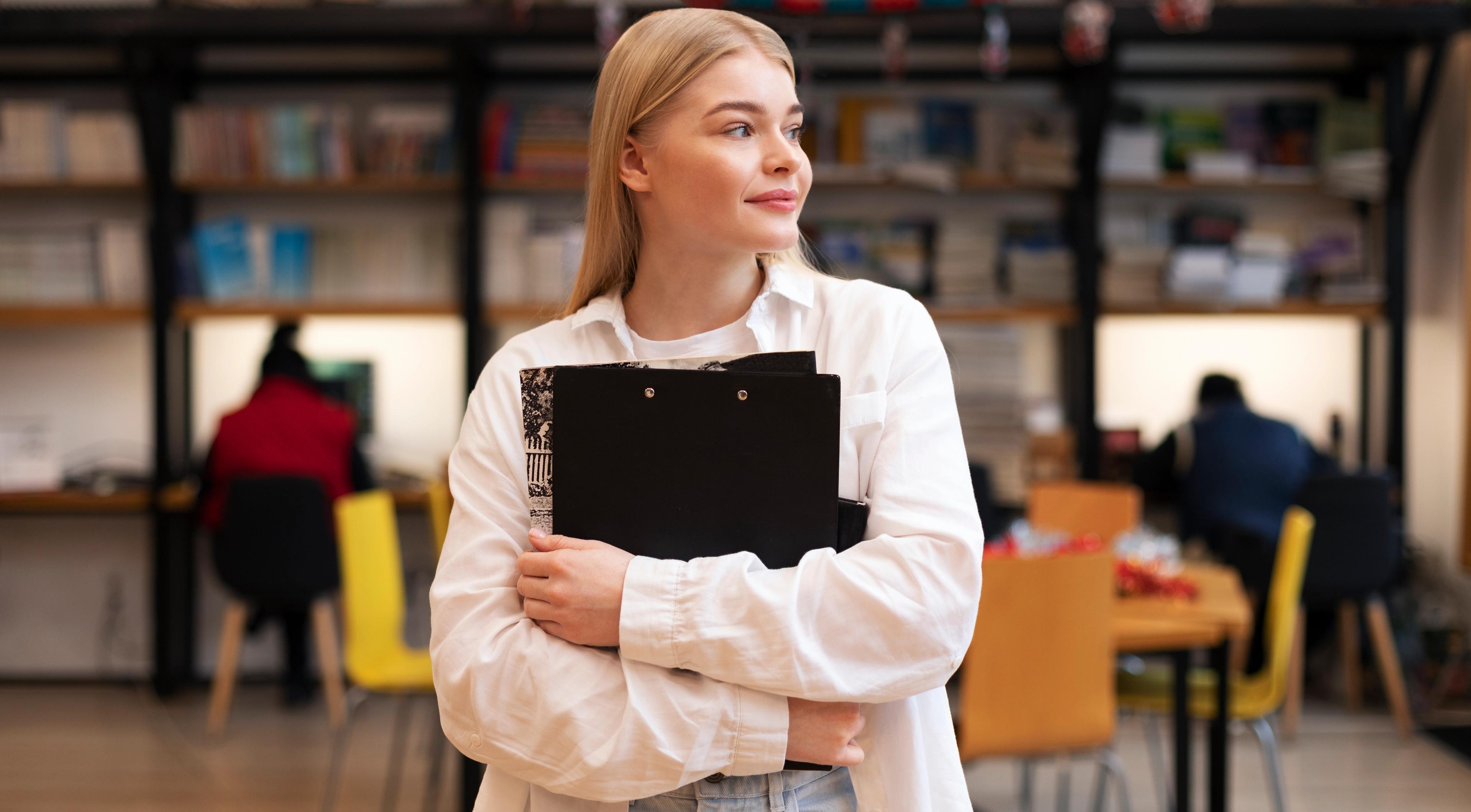 En ung kvinne står i en lesesal og holder en mappe i armene, mens hun ser tankefullt til siden.