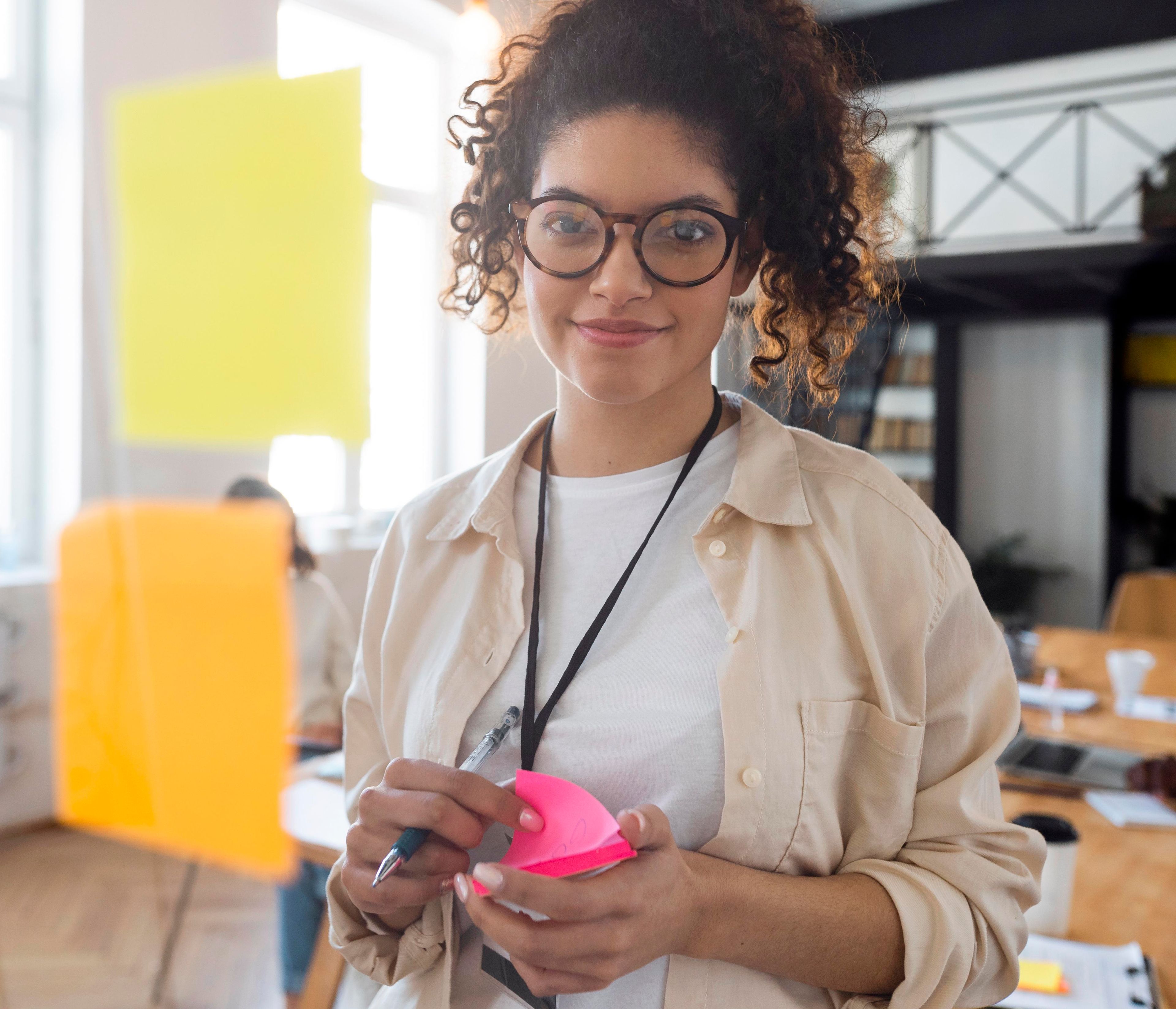 Ung kvinne med briller står i et lyst kontor og holder en rosa post-it blokk og penn foran en tavle med fargerike post it lapper.