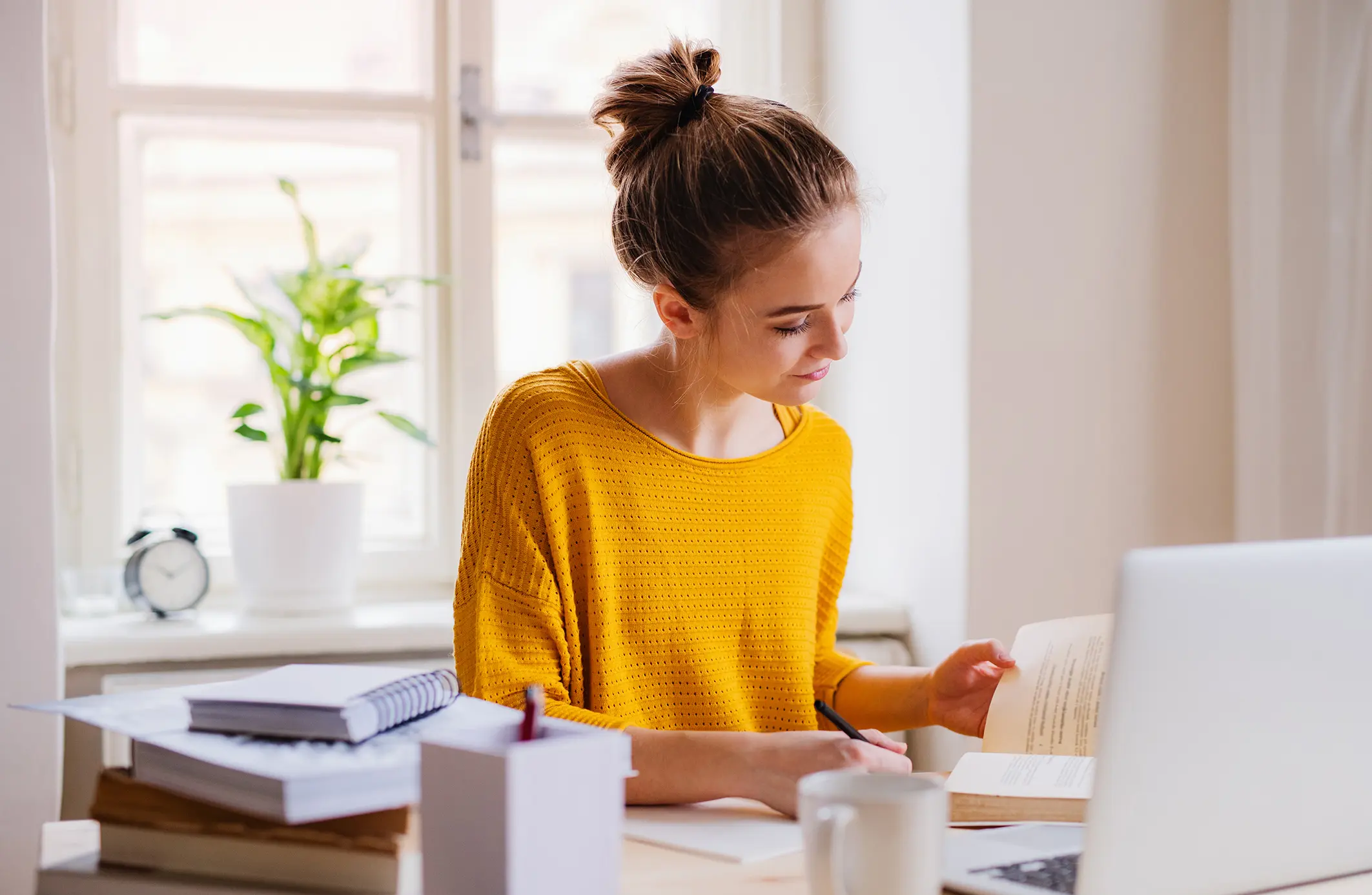 Kvinne i gul genser studerer med bøker og laptop ved et lyst vindu, omgitt av notater og kaffekopp.