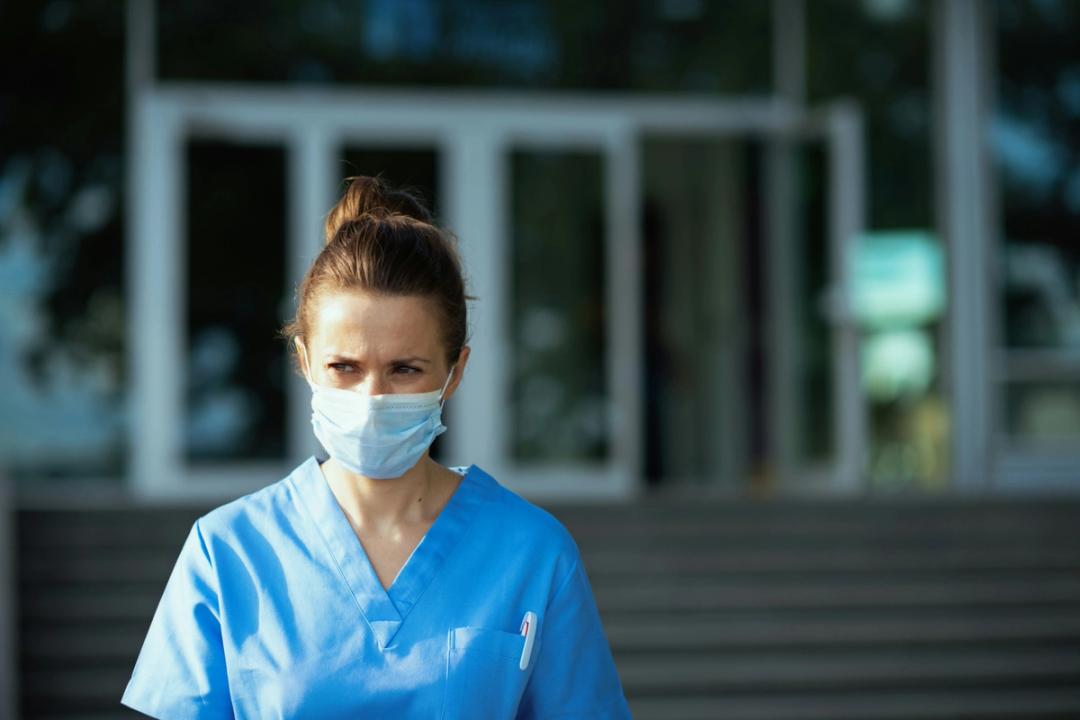 Helsearbeider i blå uniform og munnbind står utendørs foran en bygning med glassdører.