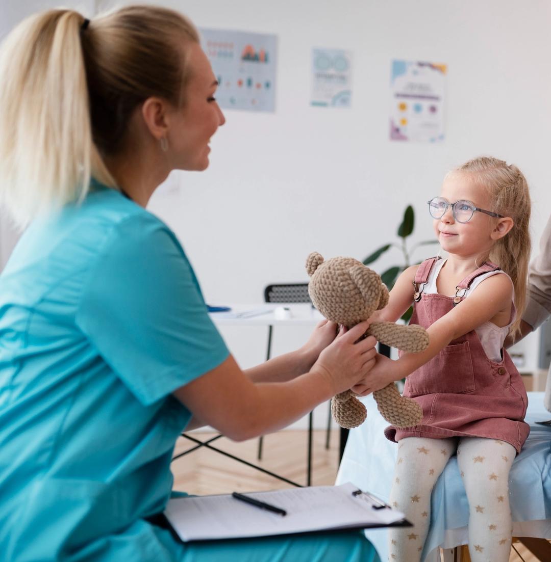 En kvinnelig helsearbeider smiler mens hun rekker en bamse til en liten jente med briller, som sitter på en benk. Jenta smiler tilbake og tar imot bamsen.