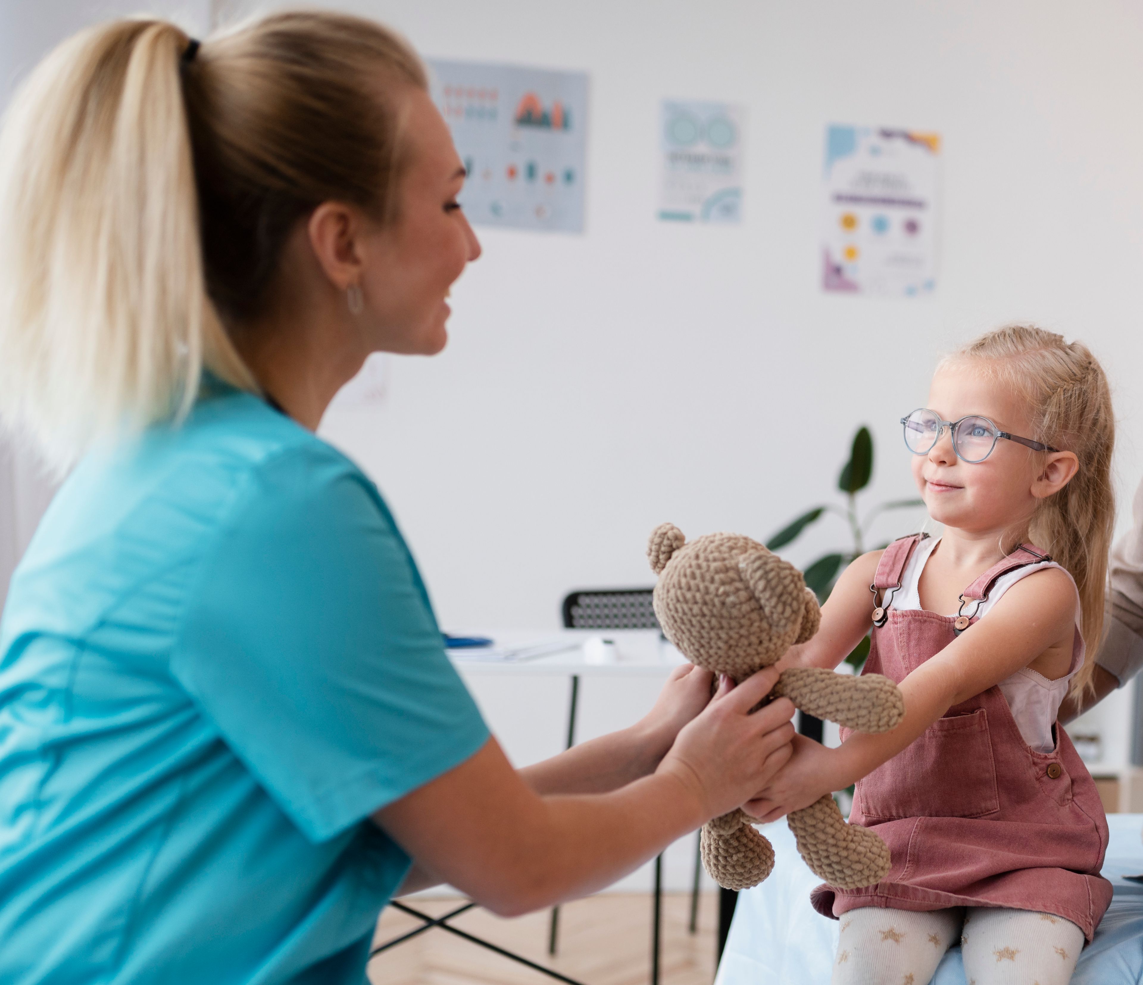 En kvinnelig helsearbeider smiler mens hun rekker en bamse til en liten jente med briller, som sitter på en benk. Jenta smiler tilbake og tar imot bamsen.