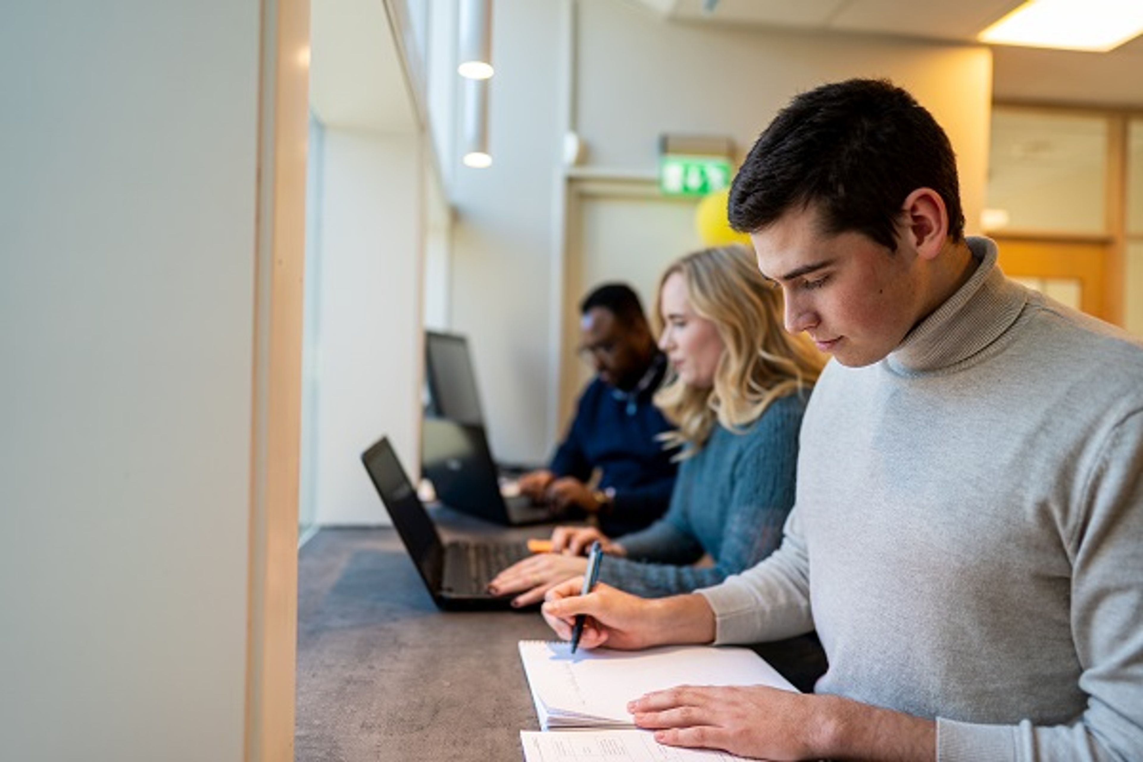 Studenter jobber ved en lang benk, noen skriver for hånd mens andre bruker laptop.