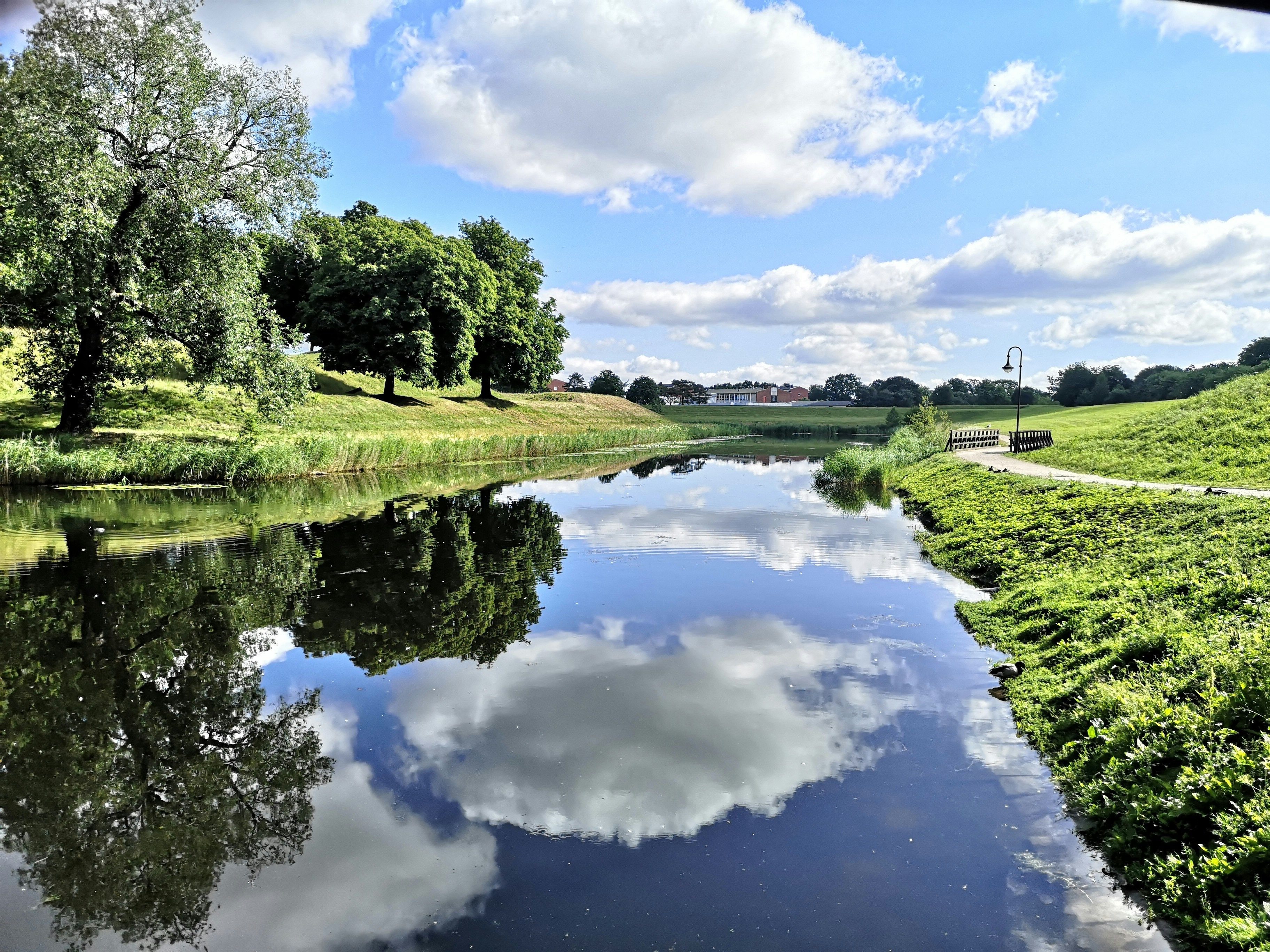 Elvepromenade i Fredrikstad med grønt gress, trær og blå himmel speilet i vannet.