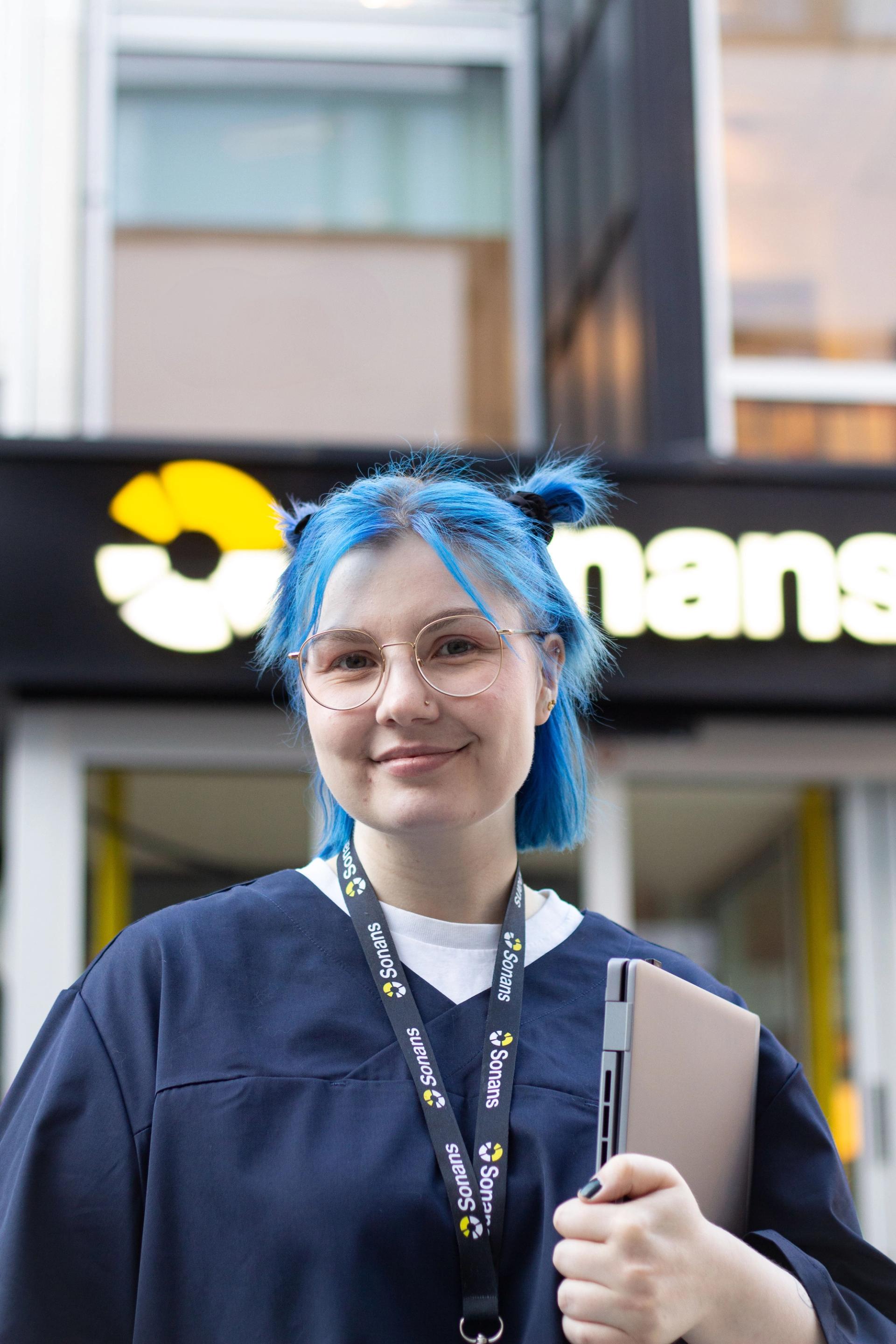 Smilende student med blått hår og Sonans-nøkkelbånd står utenfor campus med PC i hånden, klar for undervisning på yrkesfag