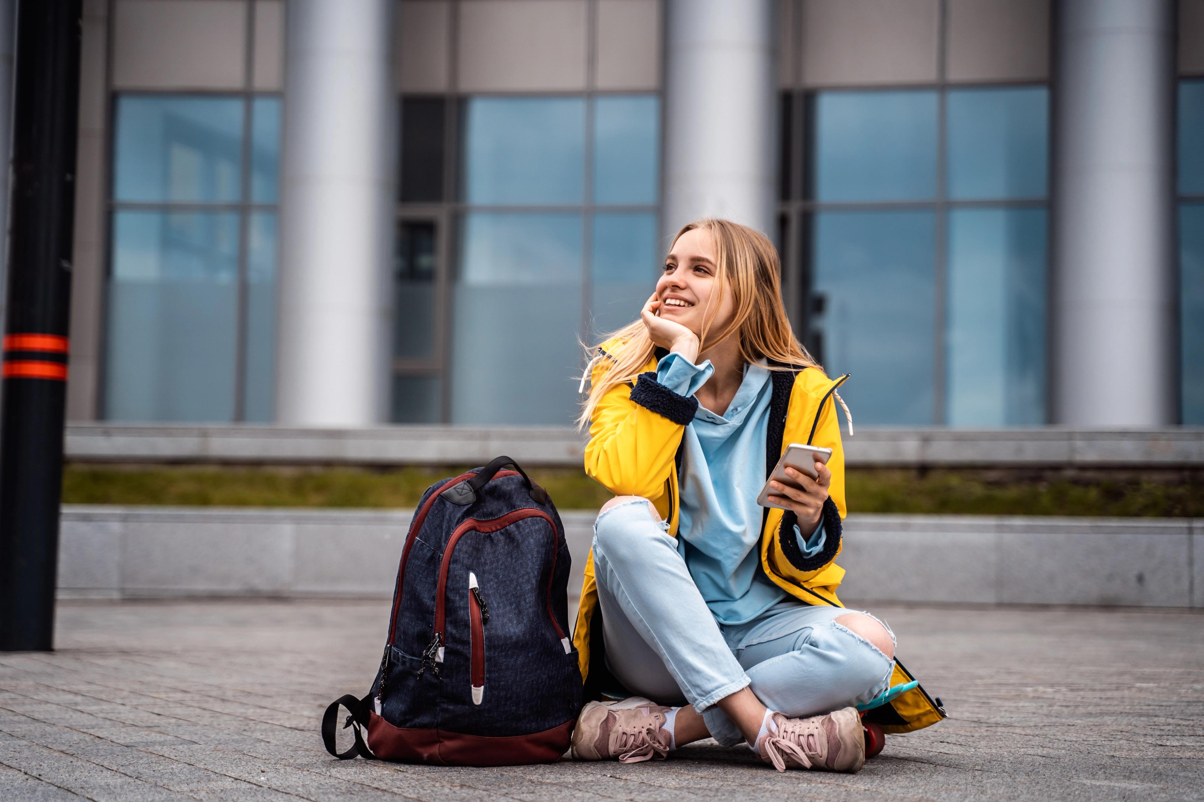 milende student med mobil og ryggsekk sitter utenfor skolebygning og ser opp - klar for en ny start som privatist.
