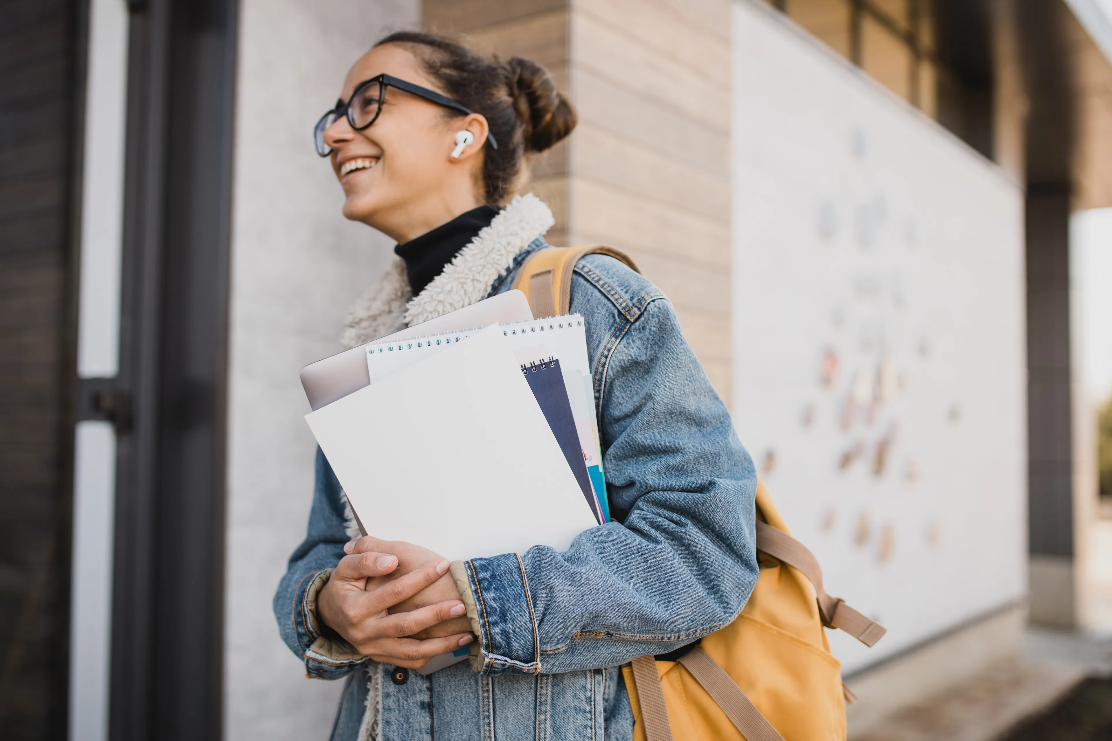 Smilende student med briller og ryggsekk holder notatbøker og laptop utendørs.