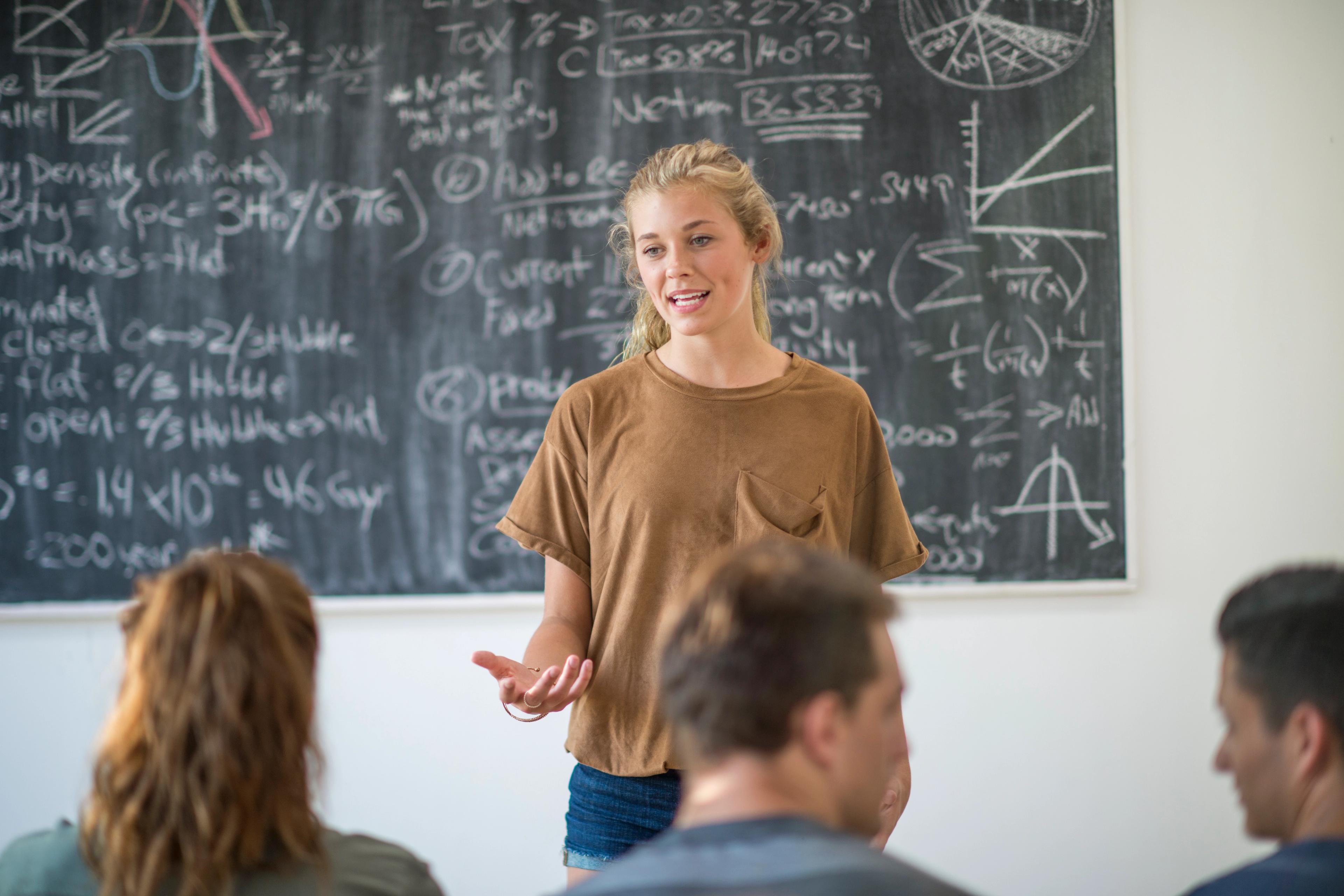 Ung kvinne holder en presentasjon foran en tavle med matematiske formler, mens medstudenter lytter.