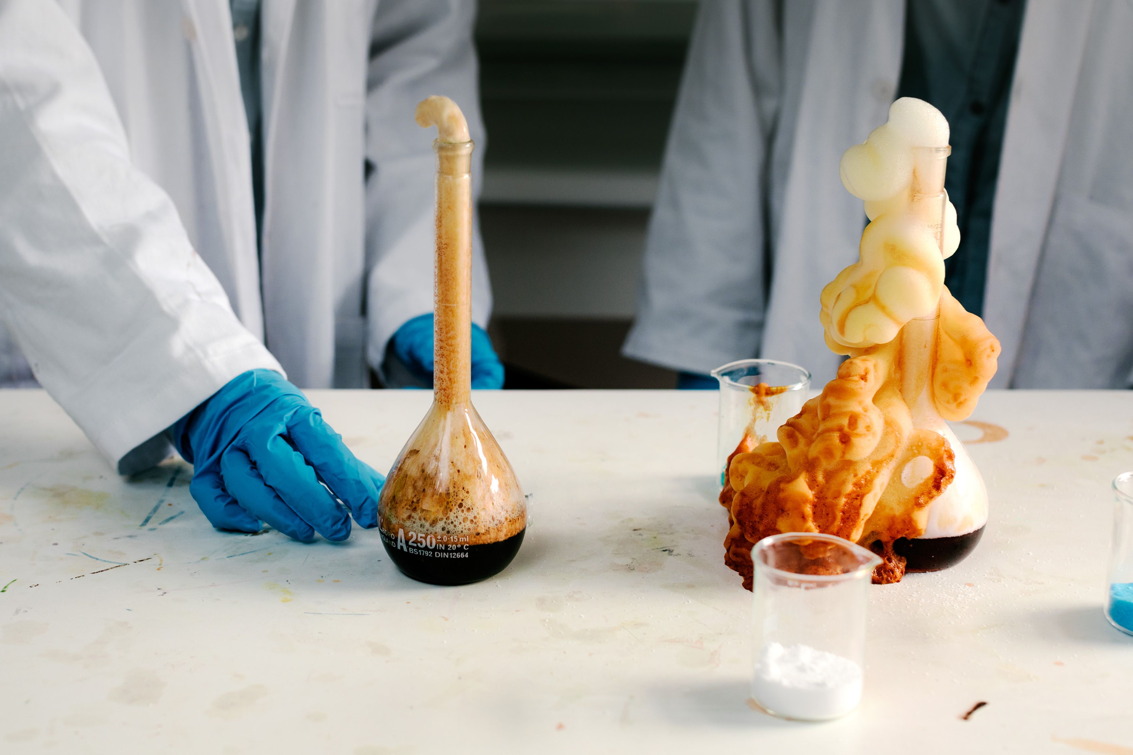 Kjemistudenter i laboratoriefrakker og hansker utfører en eksperimentell reaksjon med skum og væske.