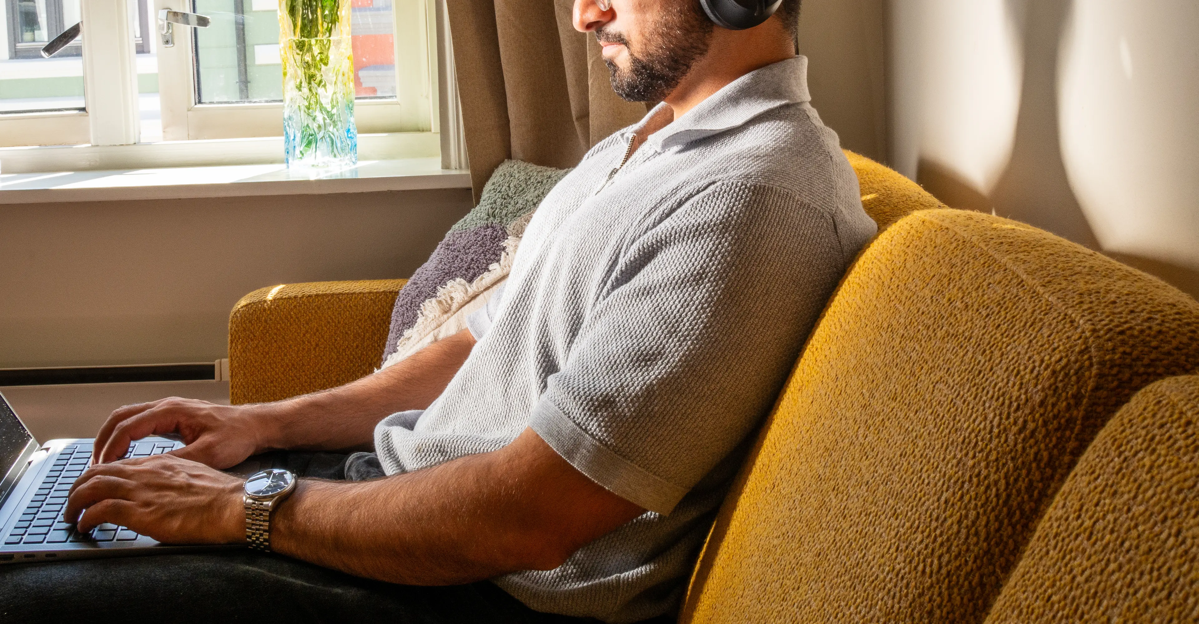 Mann med hodetelefoner jobber fokusert på laptop i solbelyst stue med blomster i bakgrunnen.
