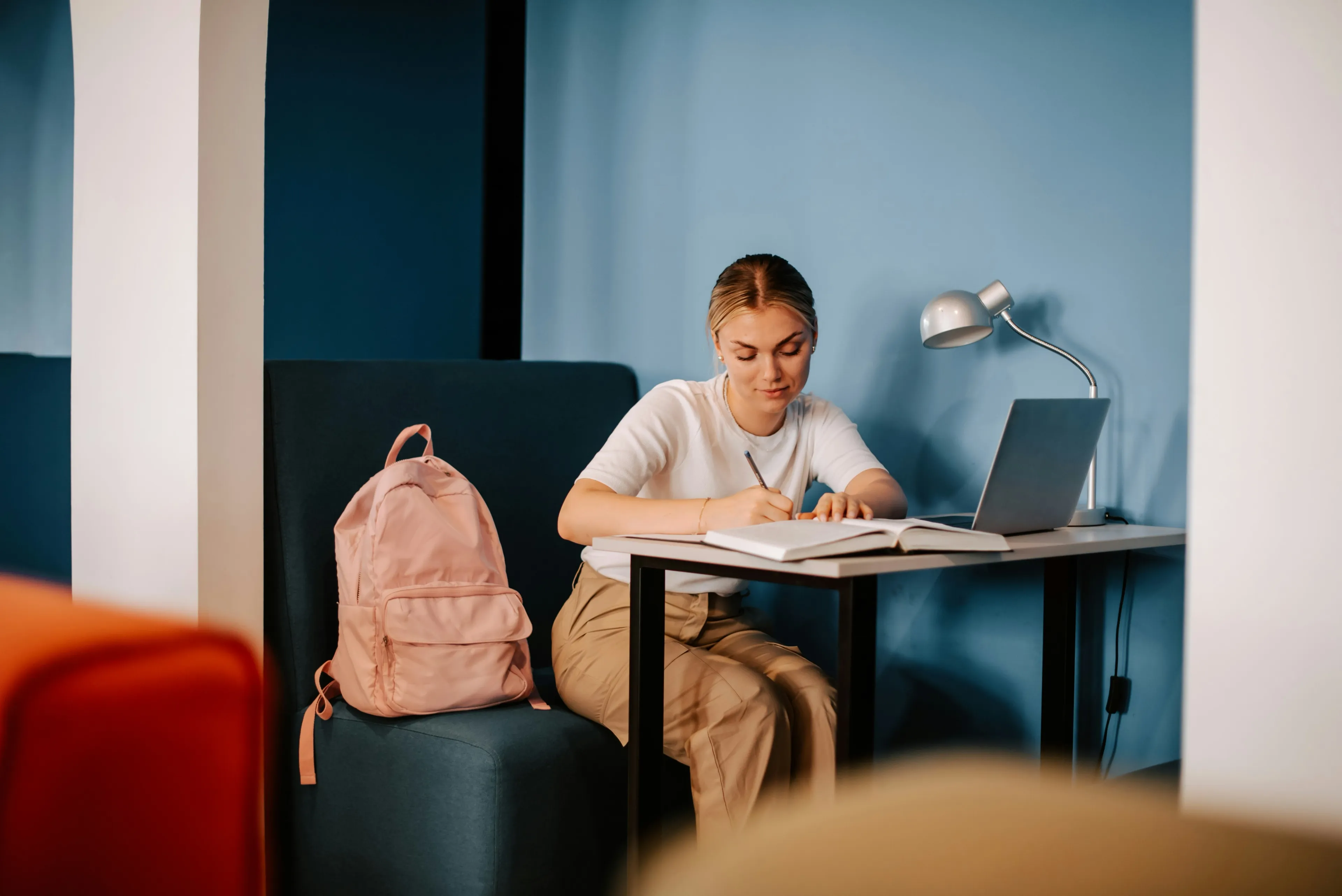 Kvinne studerer ved et skrivebord med bok, laptop og skrivebordslampe, ryggsekk ved siden av.
