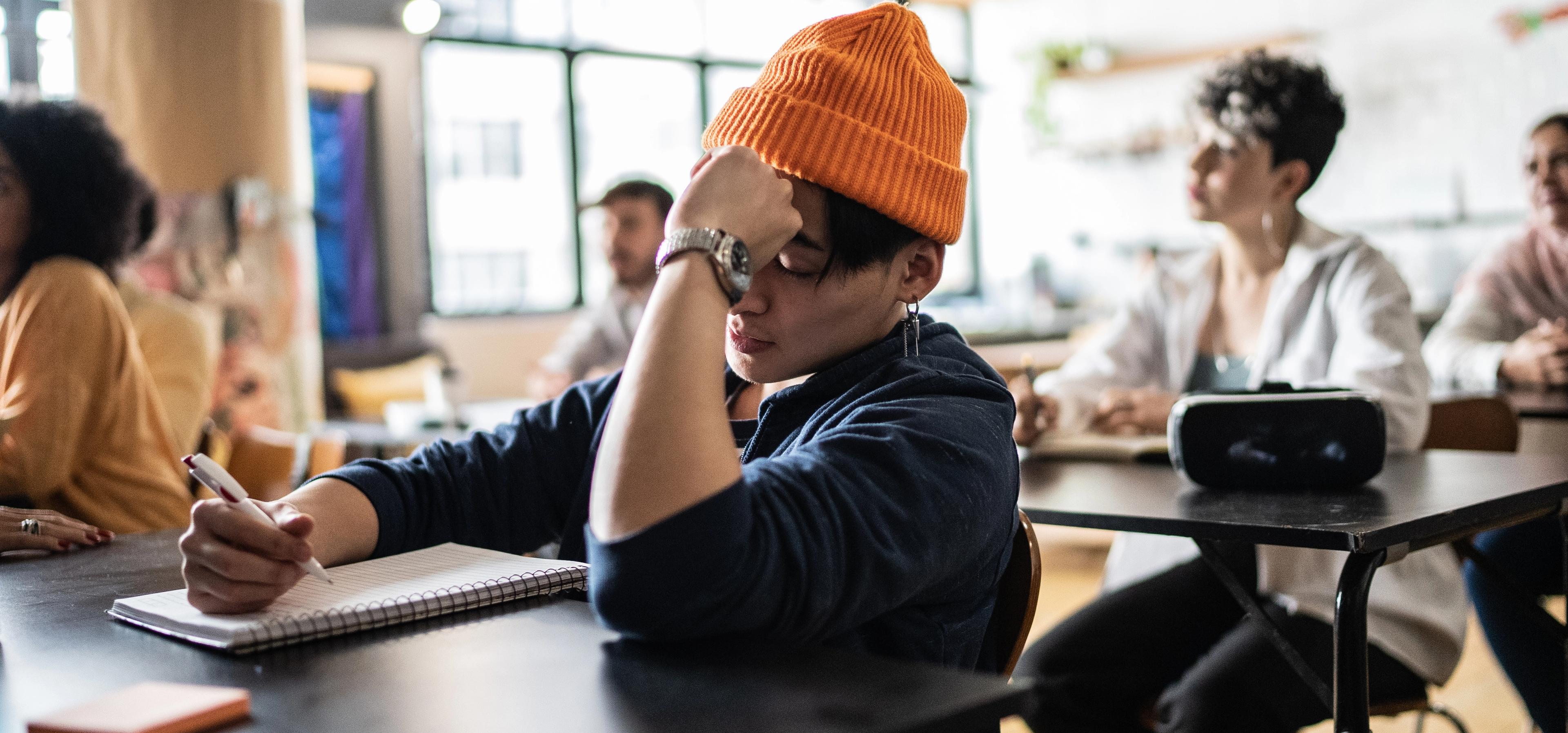Student med oransje lue sitter i et klasserom, tar notater og ser litt frustrert og umotivert ut, med hånden mot pannen. Rundt sitter andre studenter og følger med i undervisningen.