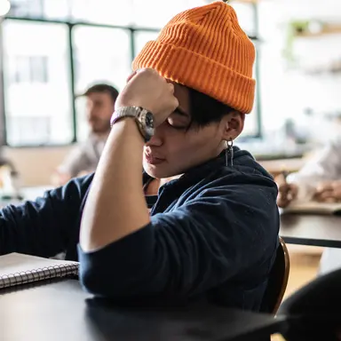 Student med oransje lue sitter i et klasserom, tar notater og ser litt frustrert og umotivert ut, med hånden mot pannen. Rundt sitter andre studenter og følger med i undervisningen.
