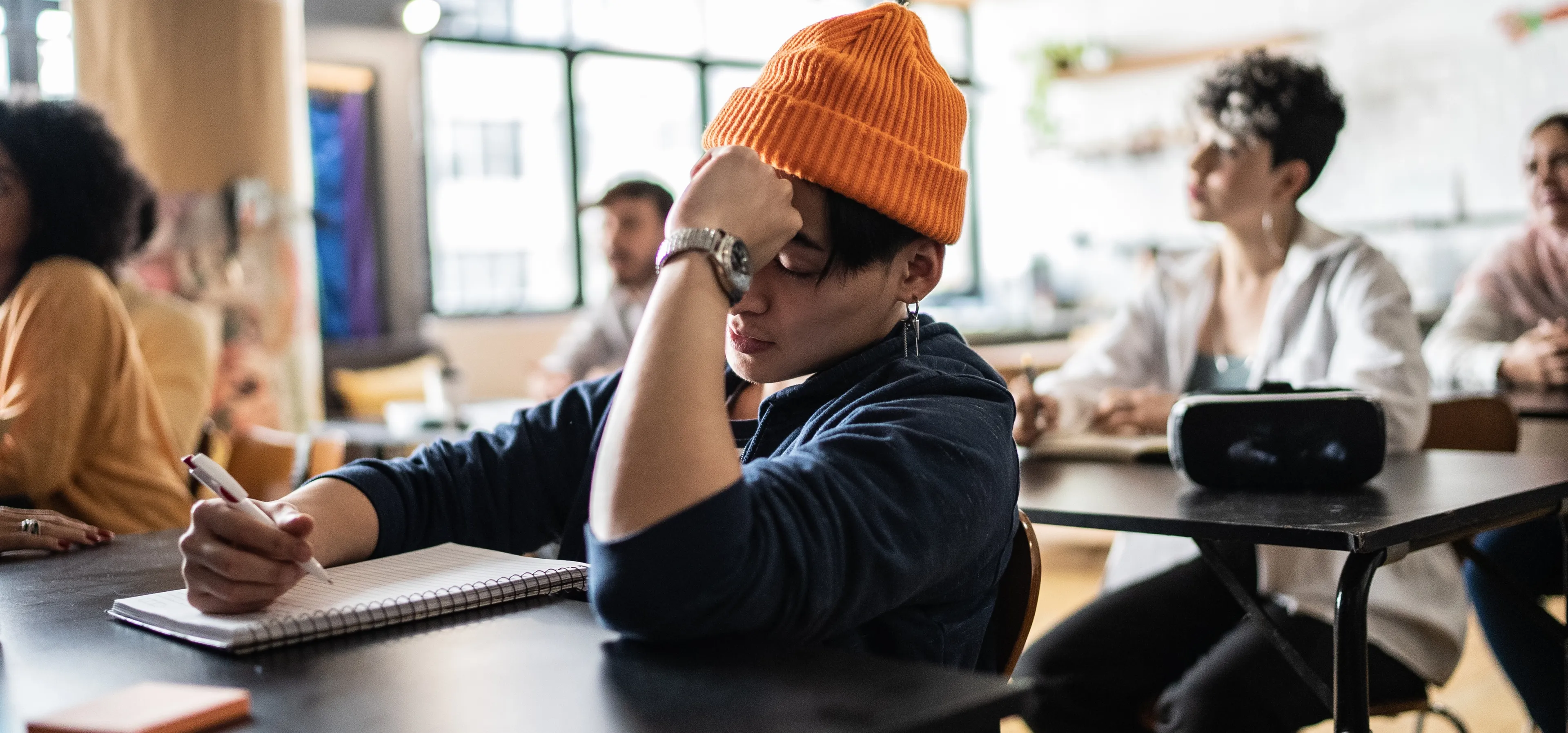 Student med oransje lue sitter i et klasserom, tar notater og ser litt frustrert og umotivert ut, med hånden mot pannen. Rundt sitter andre studenter og følger med i undervisningen.