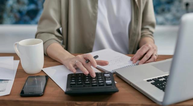 Bilde viser en person som sitter ved et bord, men en penn i hånden, regner på en kalkulator og noterer på en blokk