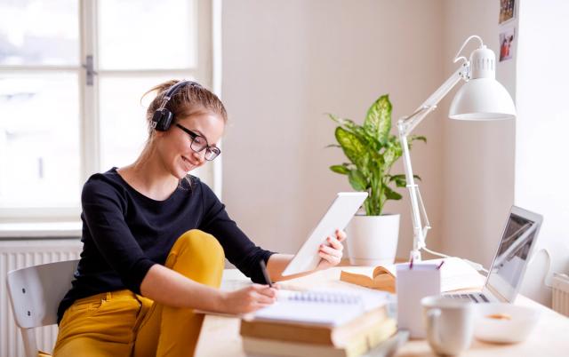 Smilende student med hodetelefoner studerer til eksamen med nettbrett, notater og laptop på en lys arbeidsplass.