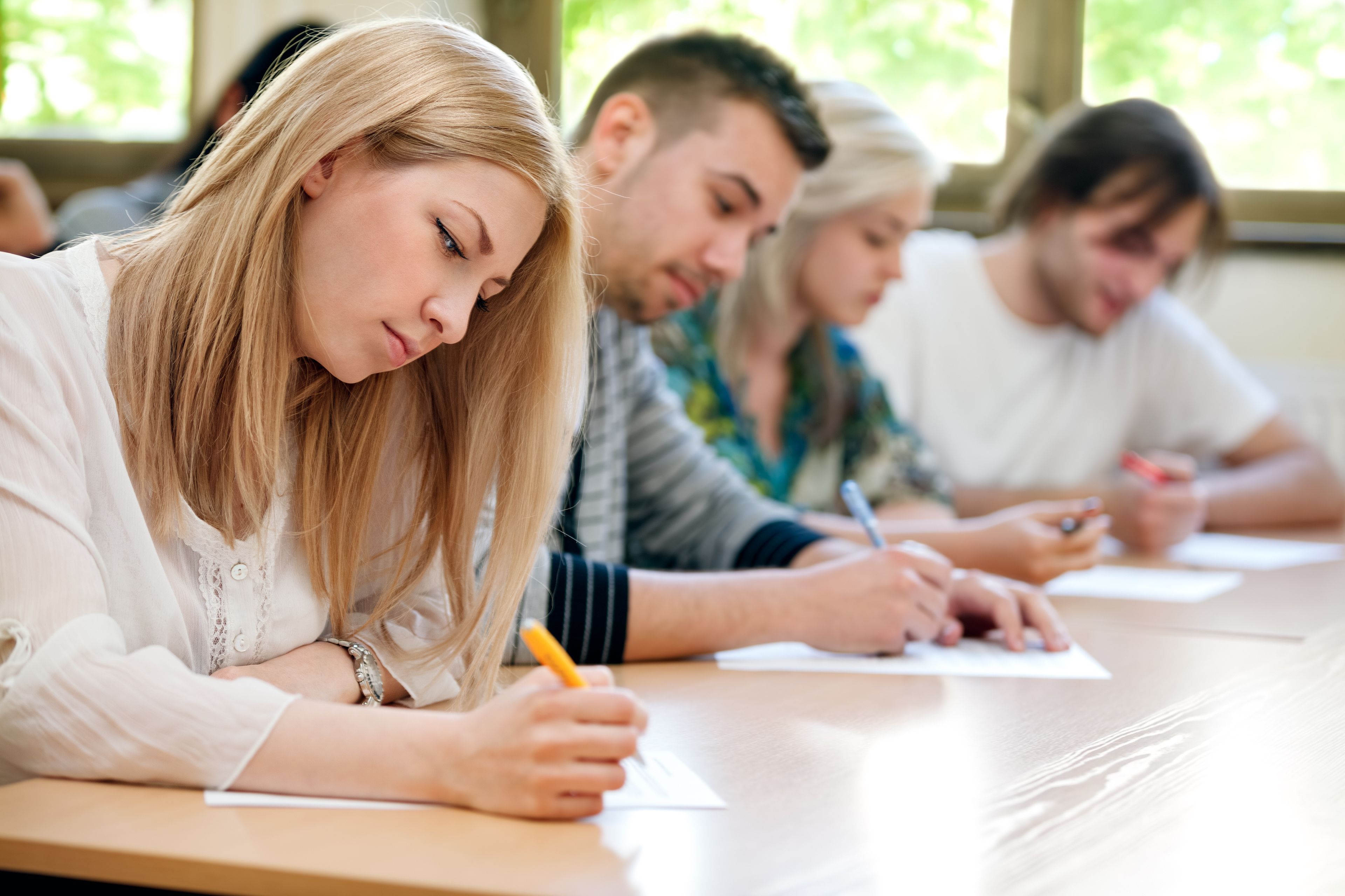 Studenter sitter ved pulter og skriver konsentrert på eksamen i et lyst klasserom.