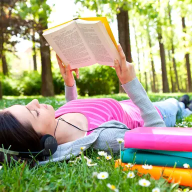 Ung kvinne leser bok ute i naturen med hodetelefoner, omgitt av skolebøker – en rolig stund for stressmestring før privatisteksamen.