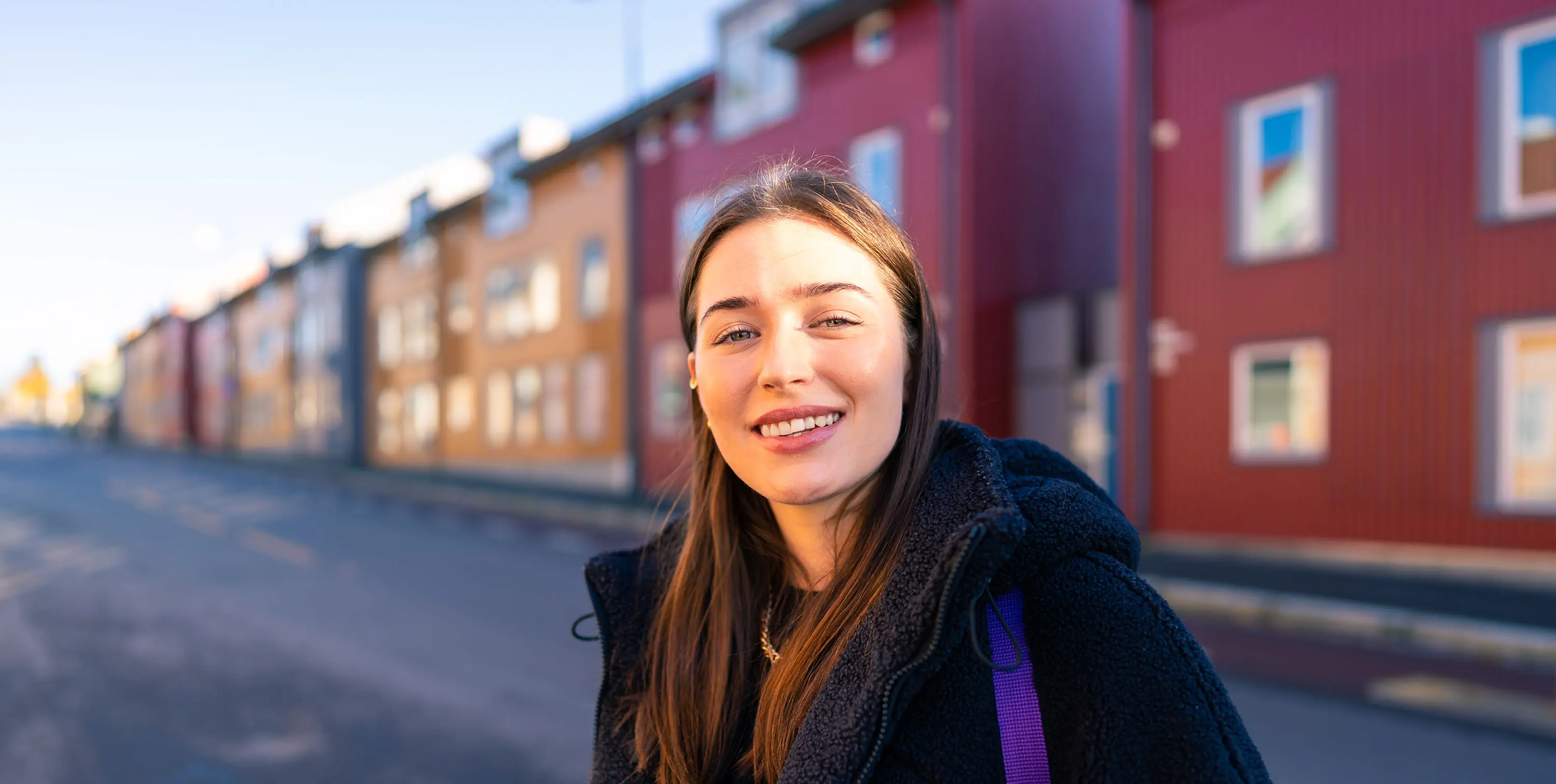 Ung kvinne smiler utendørs med fargerike bygninger i bakgrunnen på en solrik dag.