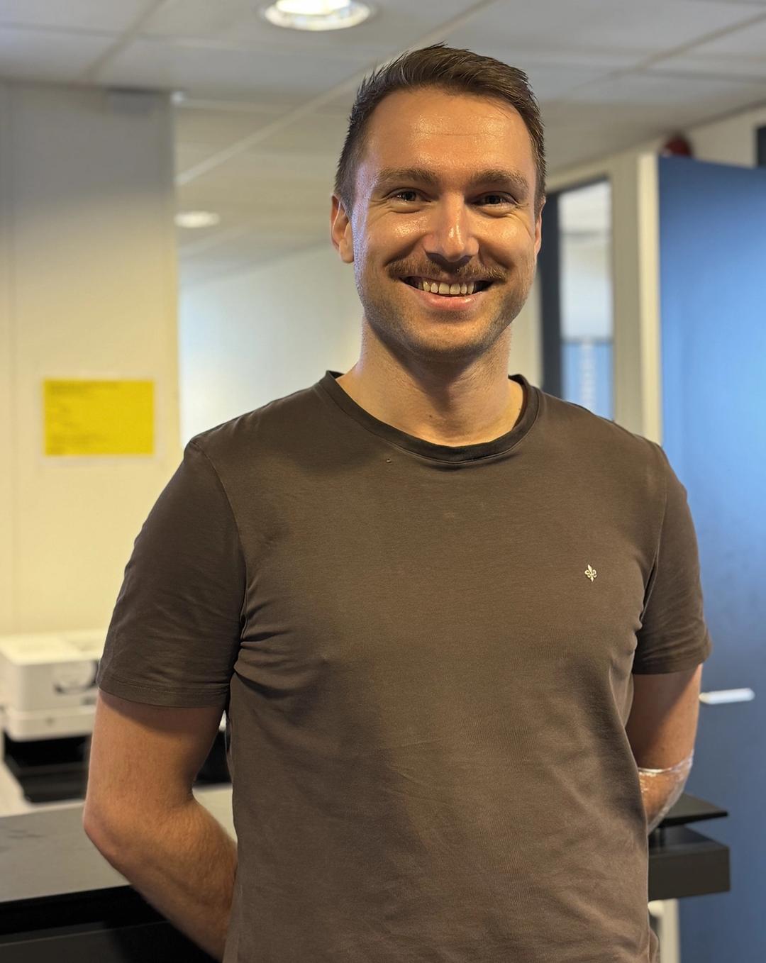 Avdelingsleder for Sonans Kristiansand, Herman Holmeide Halvorsen, smiler i et lyst atrium med gule detaljer i bakgrunnen.