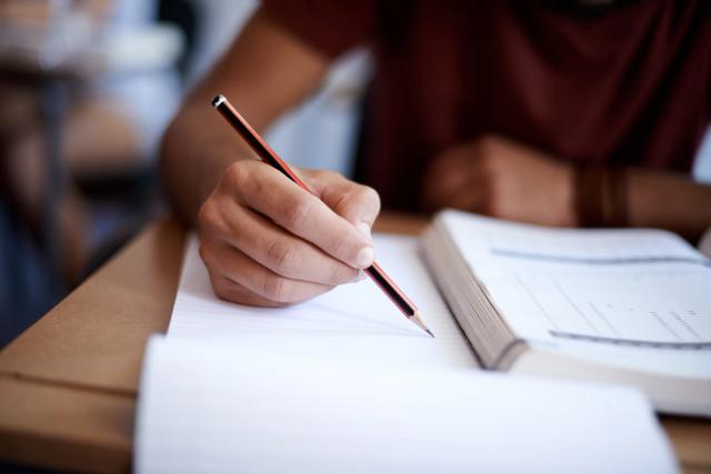 Nærbilde av en person som skriver notater for eksamensforberedelse, med bok og skriveblokk på pulten.