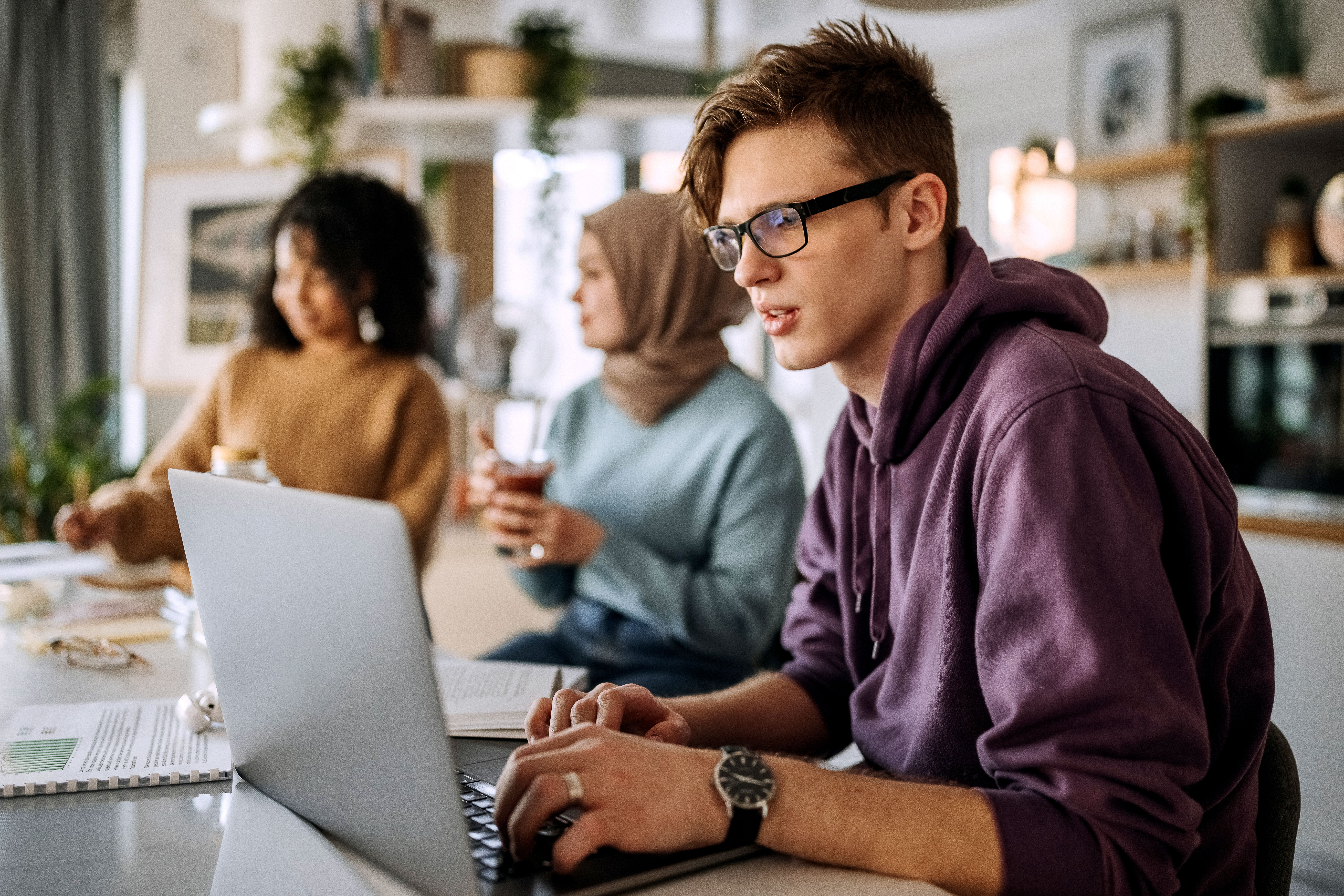 Tre studenter jobber sammen ved et bord. En mann bruker PC, mens to kvinner snakker i bakgrunnen.