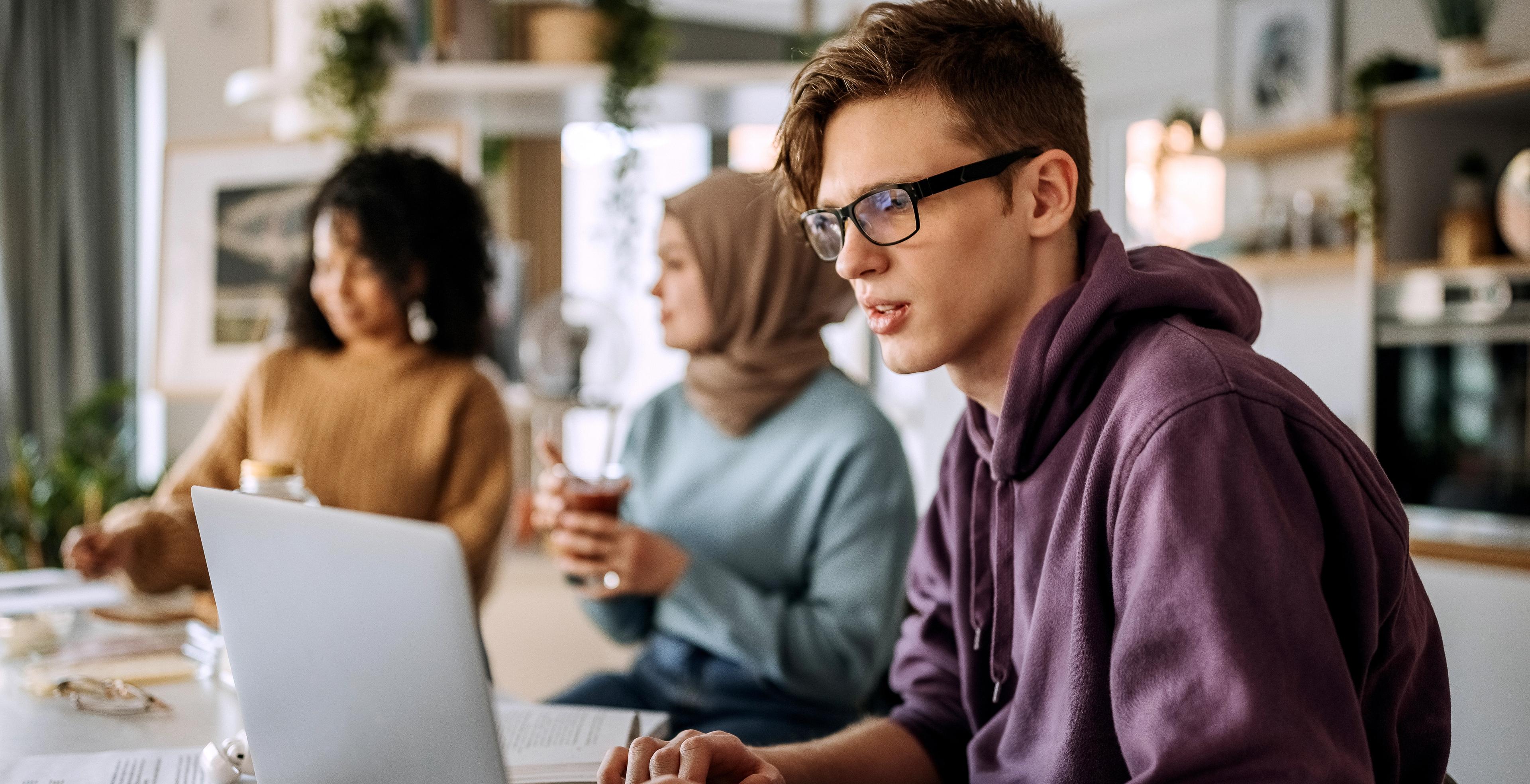 Tre studenter jobber sammen ved et bord. En mann bruker PC, mens to kvinner snakker i bakgrunnen.