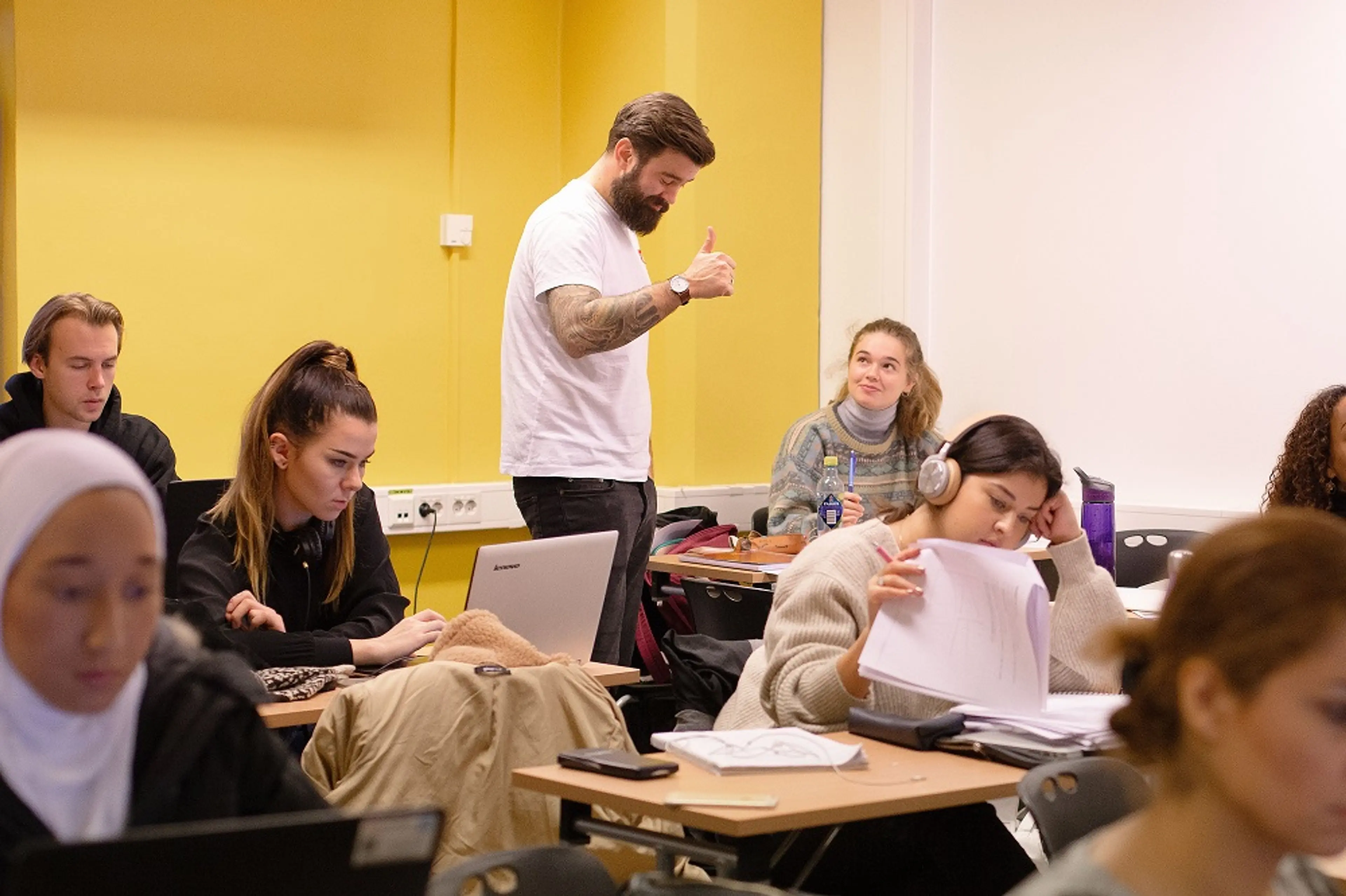 Studenter i klasserom på under forelesning. En lærer gir tommel opp mens elevene jobber med oppgaver på PC og papir. Miljøet er fokusert og engasjerende.