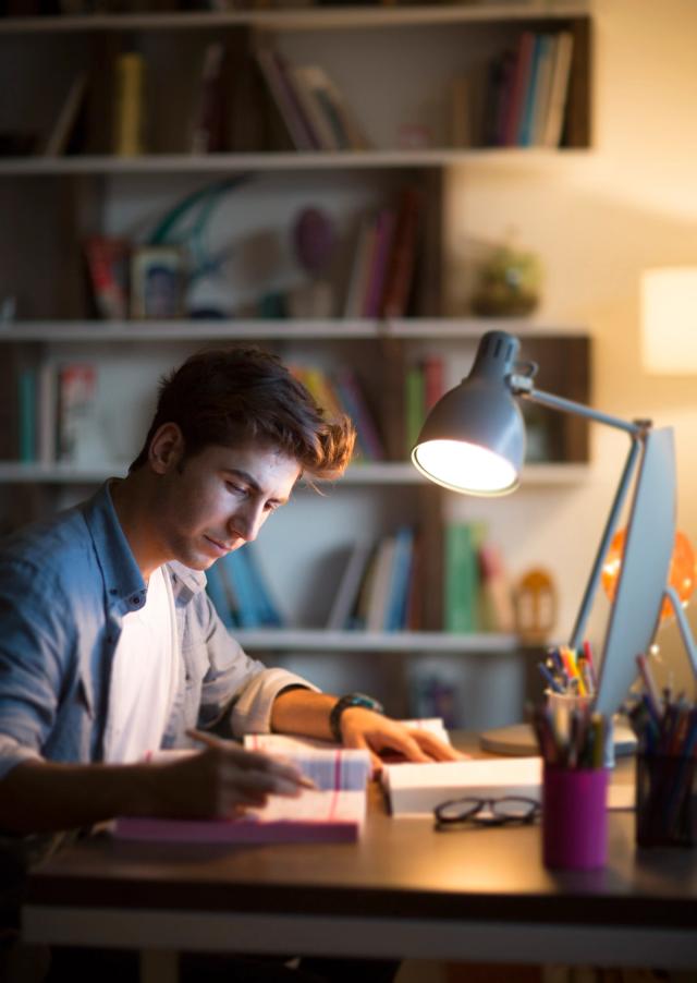 Ung mann studerer til eksamen ved et skrivebord med bøker og PC, belyst av en skrivebordslampe.