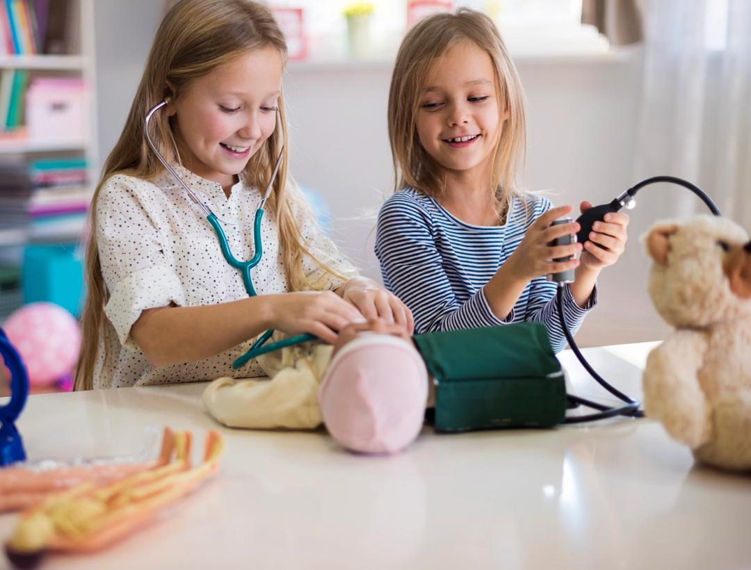 To små jenter leker doktor med dukken og bamsen sin, de lytter med stetoskop og måler blodtrykk.