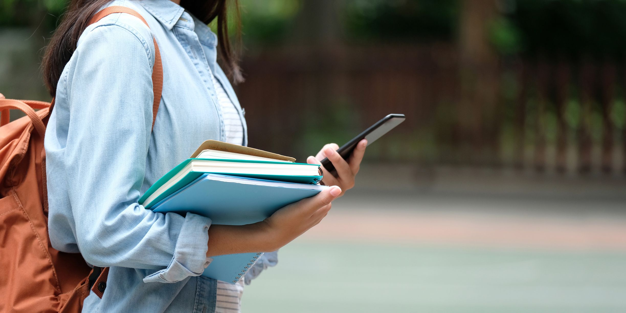 Student med ryggsekk holder bøker og mobiltelefon mens hun går utendørs.