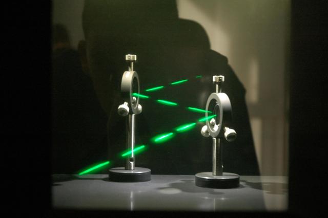 Et grønt laserlys sendes gjennom et optisk oppsett med to komponenter montert på stativer. Strålen passerer gjennom åpninger og blir delt i flere retninger, noe som tyder på interferens eller diffraksjon. Dette er et eksempel på hvordan lys oppfører seg som en bølge, der bølgelengde og fase bestemmer mønsteret som dannes. Eksperimentet brukes i fysikk for å studere lys og dets egenskaper.