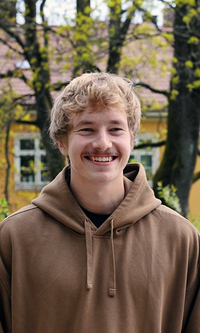 En ung mann med bart og brun genser smiler. Han står foran et gult hus og grønne trær.