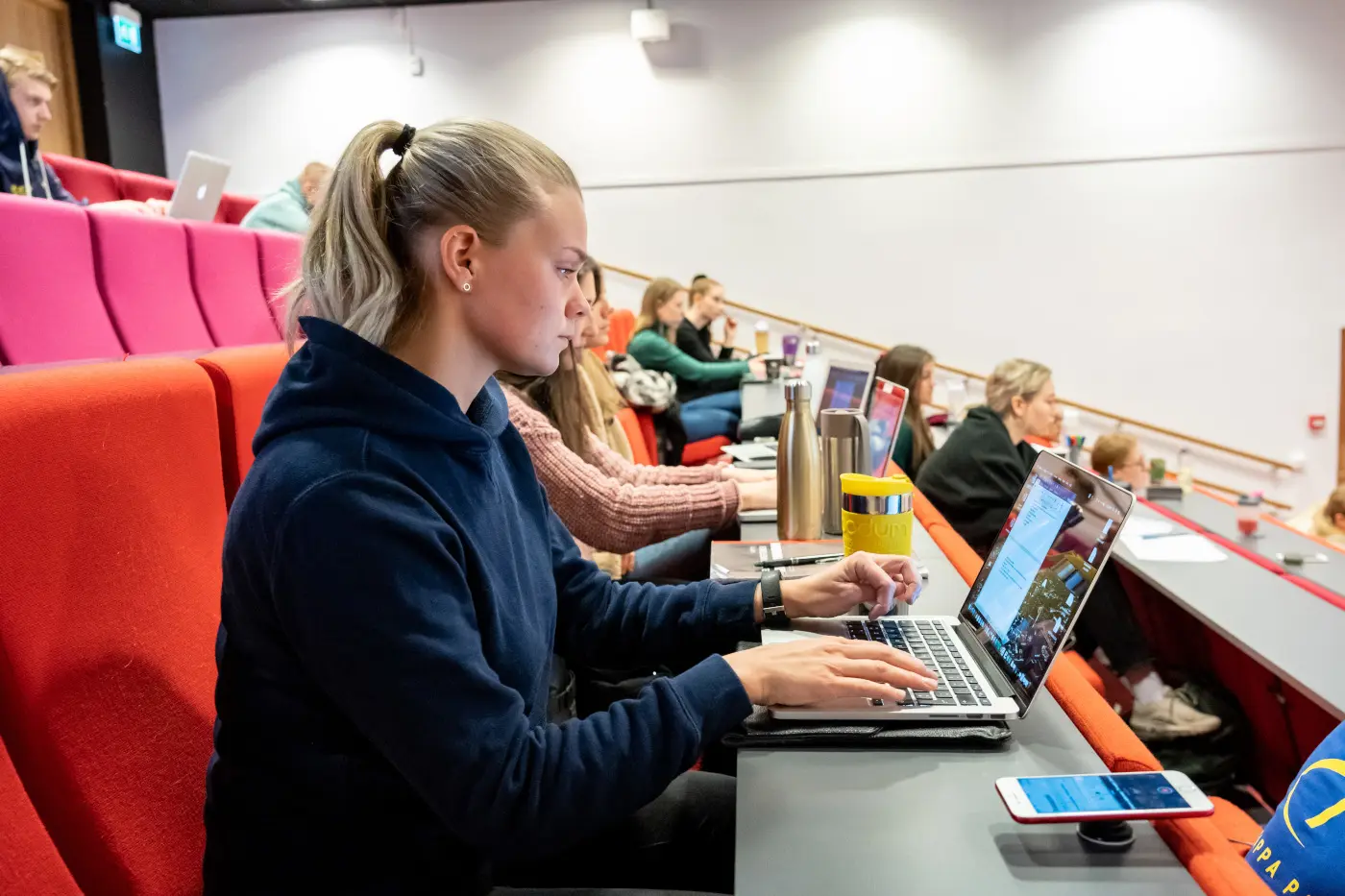 Student tar notater på laptop i en forelesningssal, omgitt av medstudenter med bøker og kaffekopper.