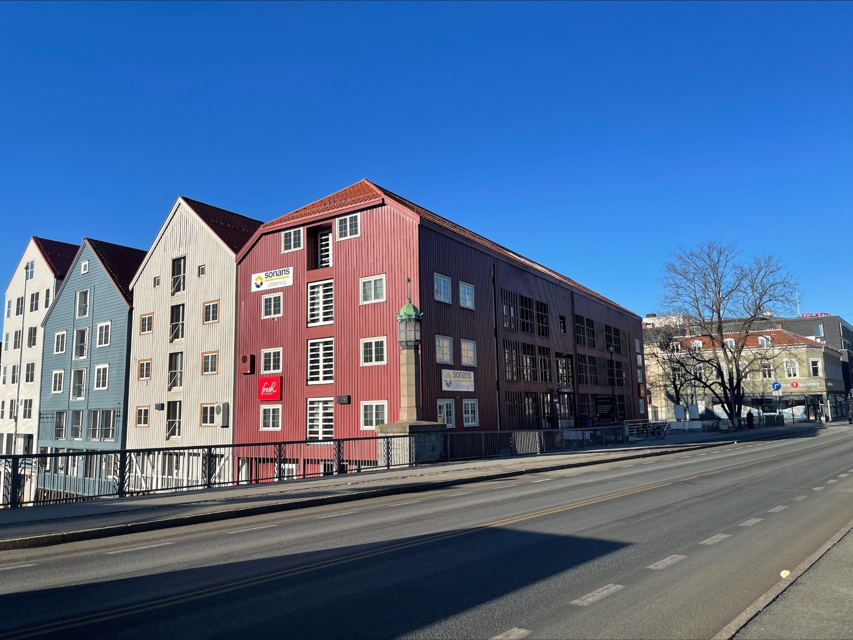 Alt-tekst: Fasaden til Sonans Trondheim, et rødt trebygg langs veien, omgitt av fargerike bryggehus.