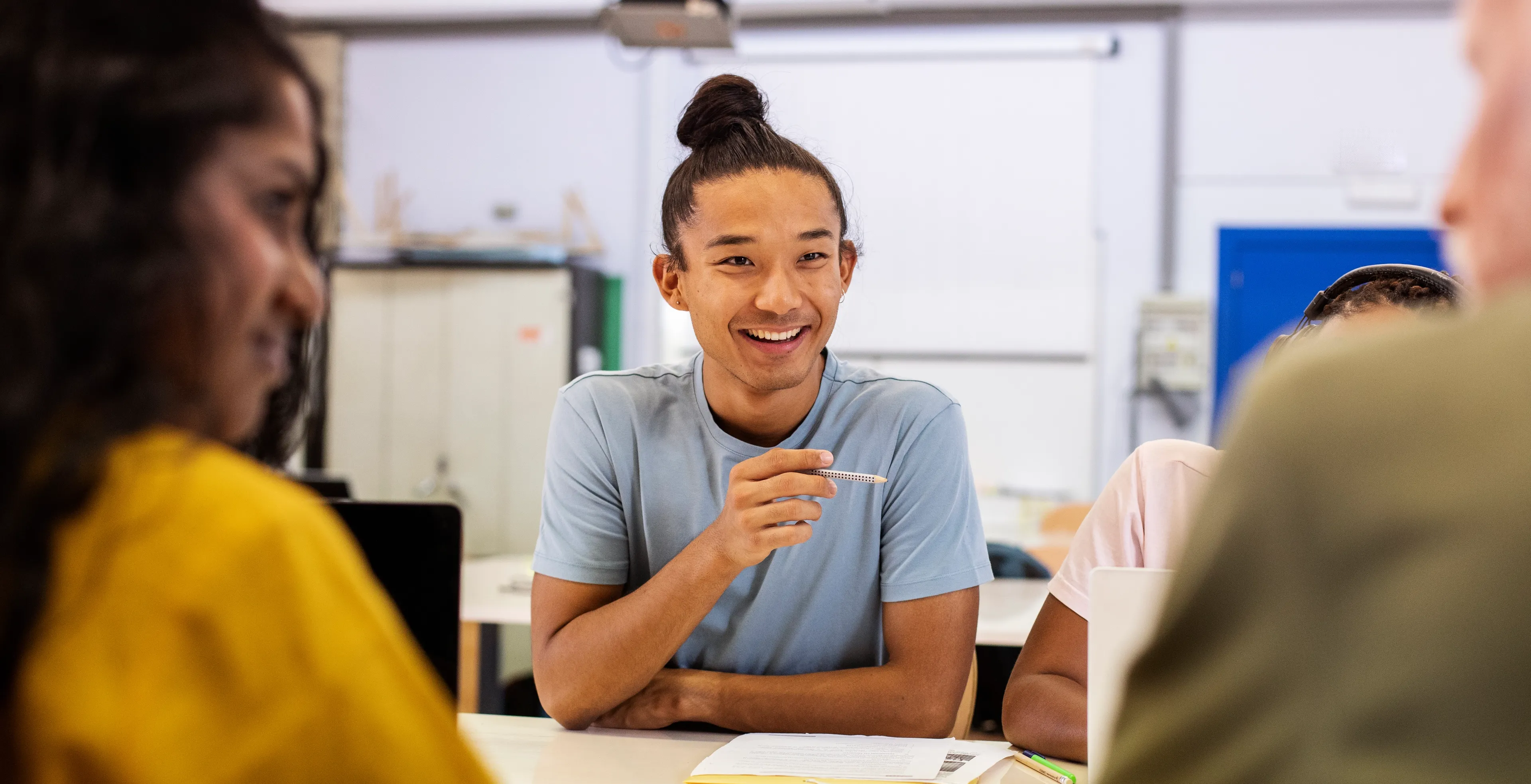 En smilende student diskuterer med andre rundt et bord i et lyst klasserom.