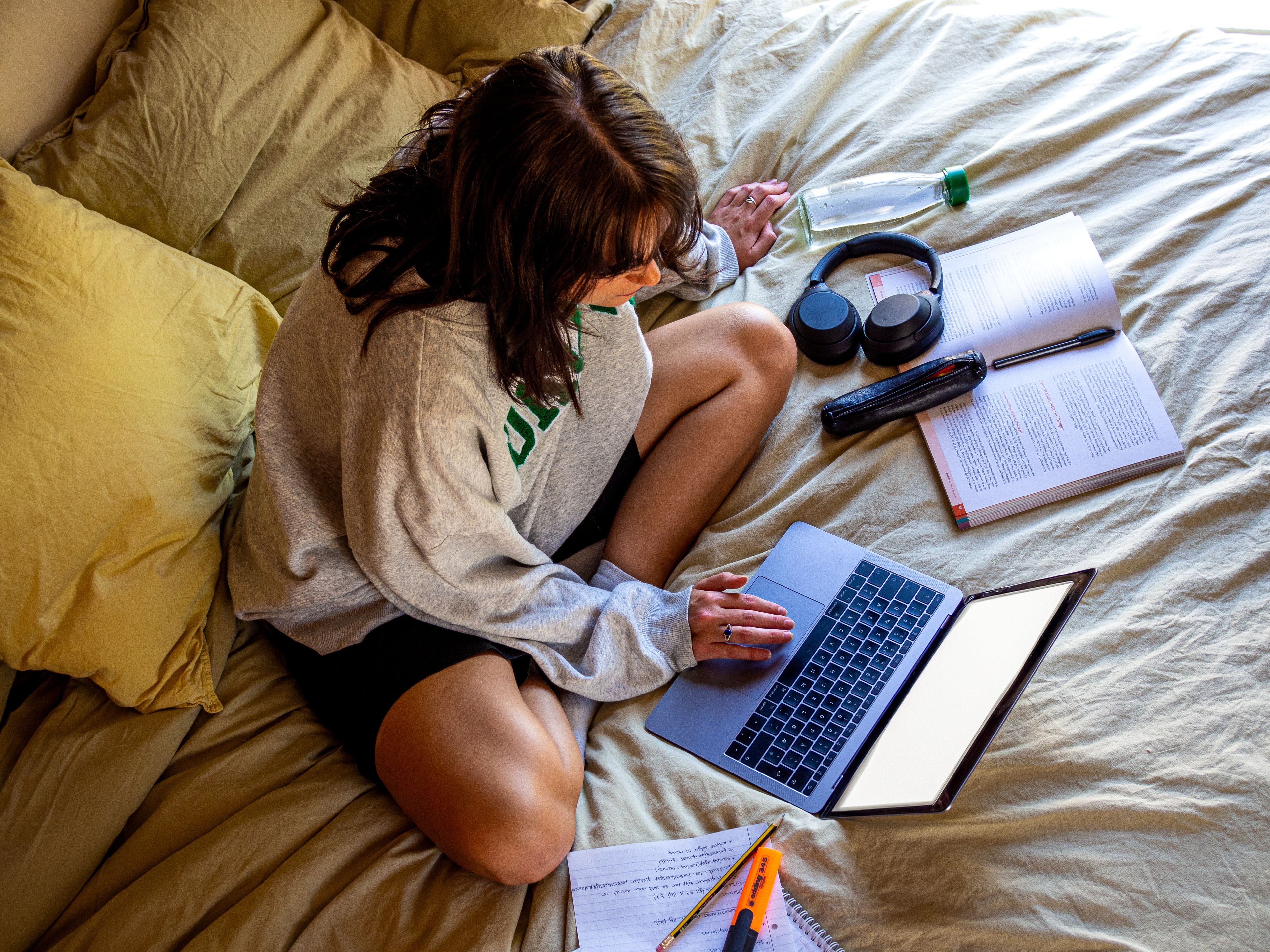 Student jobber på laptop på sengen, omgitt av notater, lærebok, hodetelefoner og vannflaske.