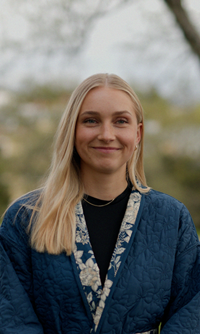 Smilende ung kvinne i blå jakke foran grønn, naturlig bakgrunn.
