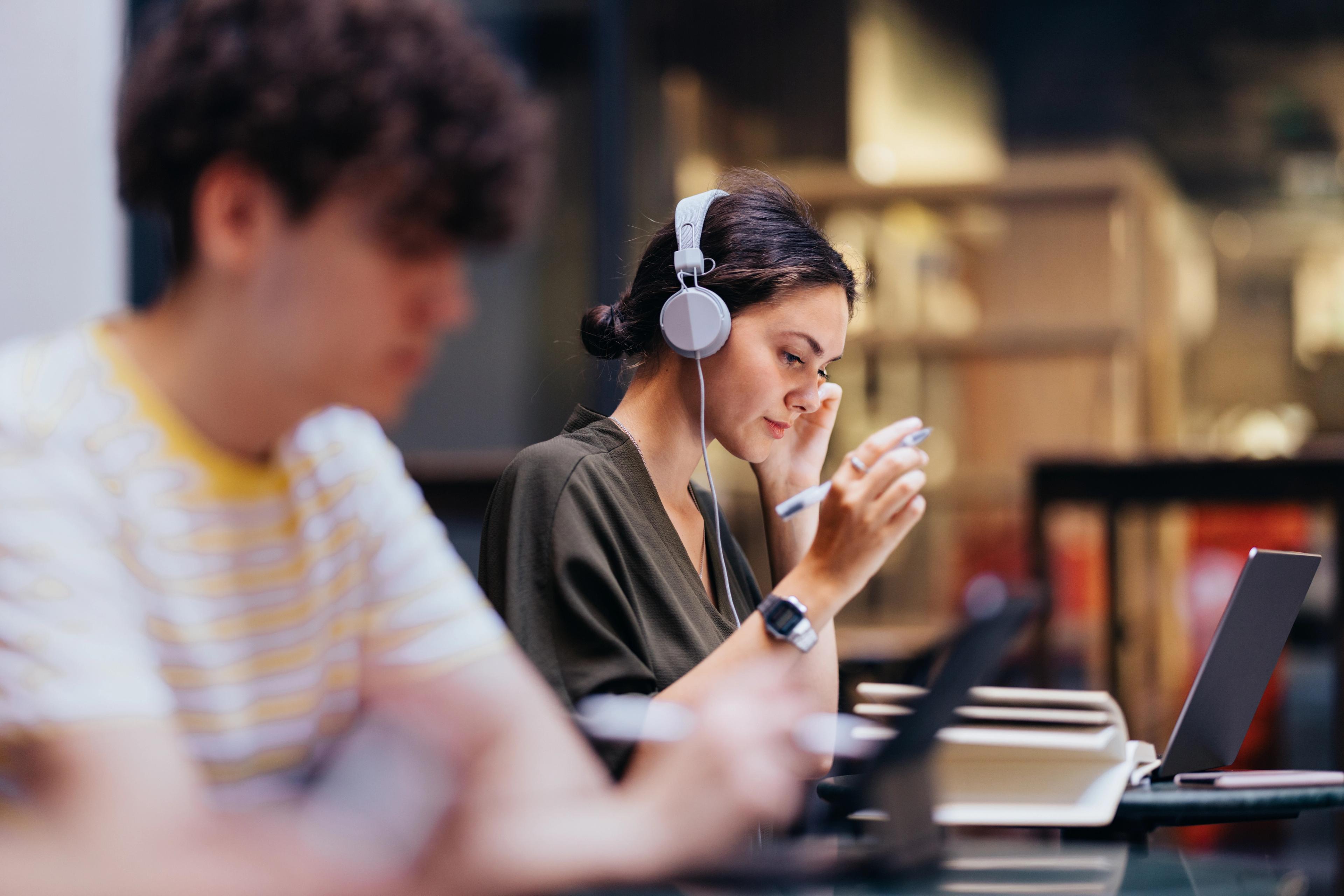 To personer studerer. Jenta, i fokus, har hodetelefoner, penn og laptop, og ser konsentrert ut.Student ved Sonans leser i en lærebok ved en studieplass med avskjermede båser og en gul vegg i bakgrunnen.