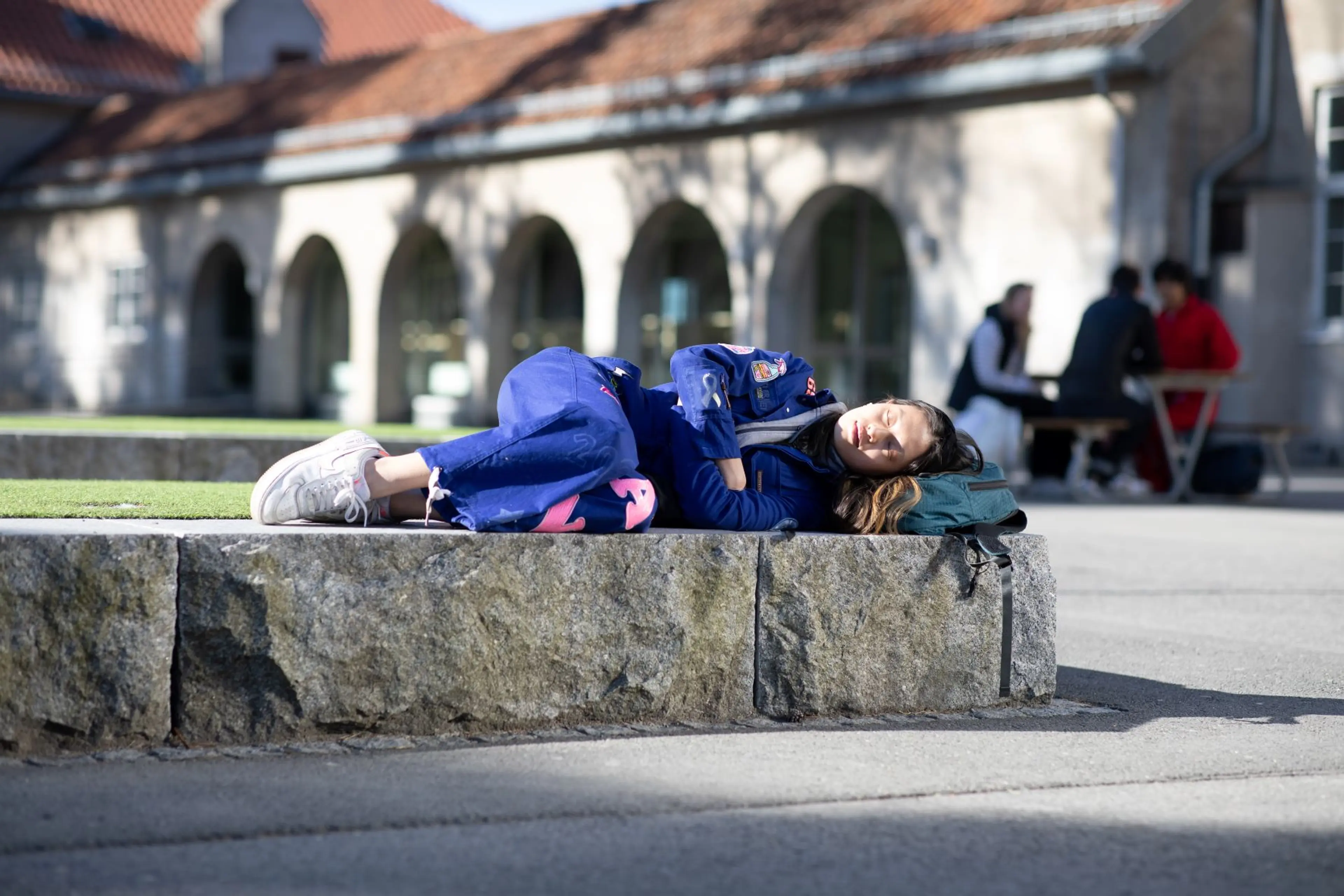 Student i blå russedress hviler på en steinbenk i solskinn, med en ryggsekk som hodepute.