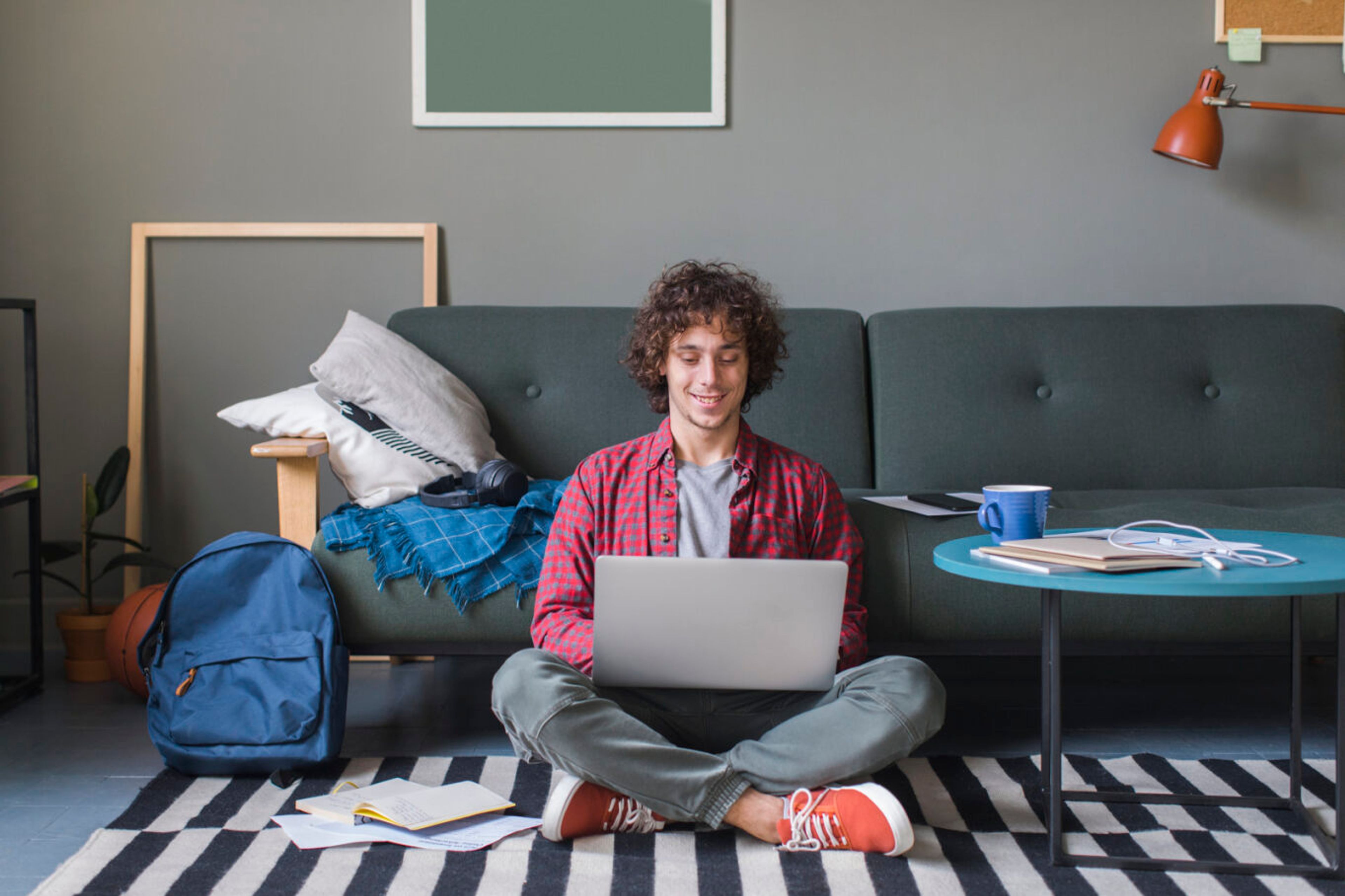 Smilende ung mann med laptop sitter på gulvet i en stue med sofa, bøker og ryggsekk.