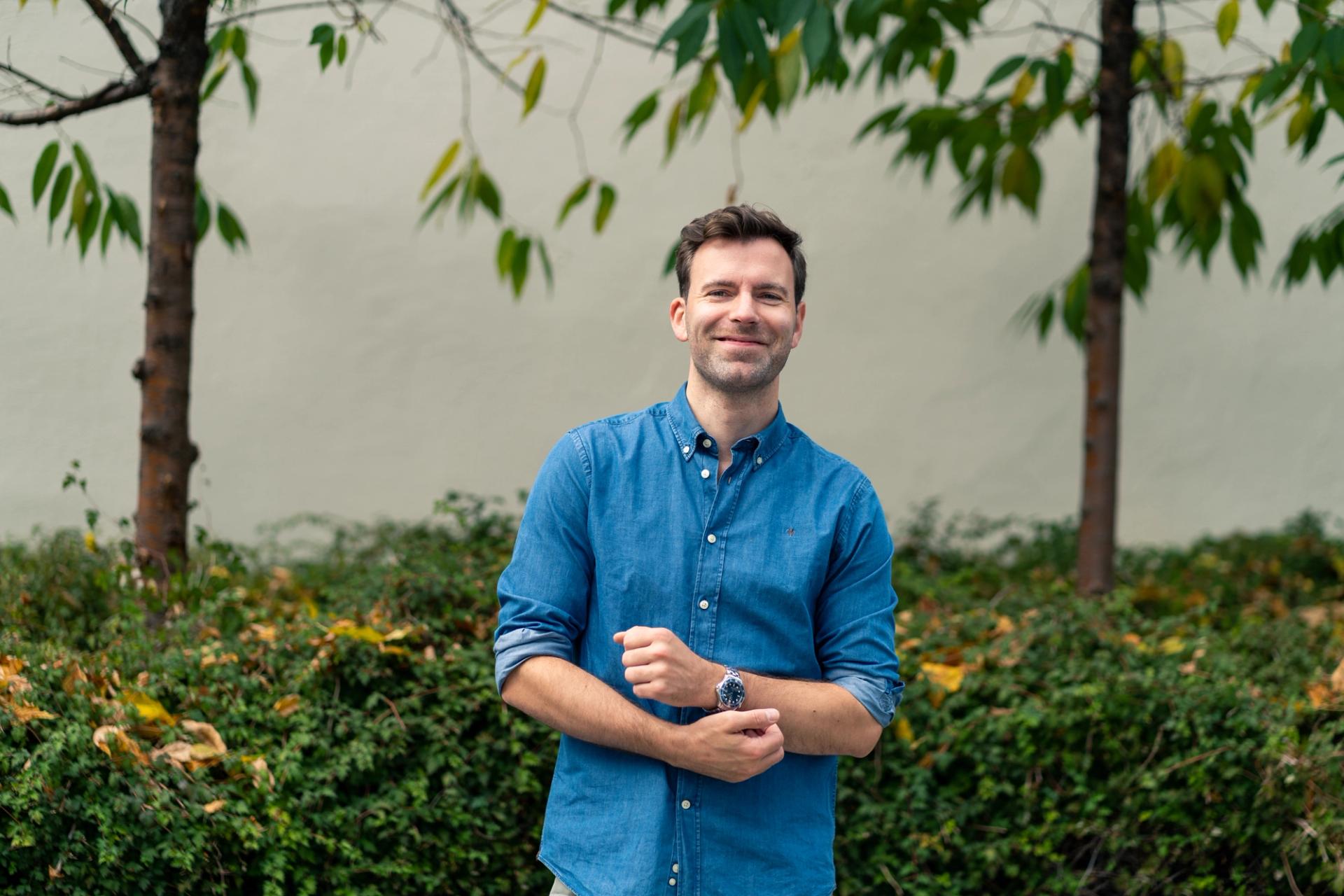 Lærer Andreas Stormo smiler og står utendørs foran grønn beplantning og en lys vegg.
