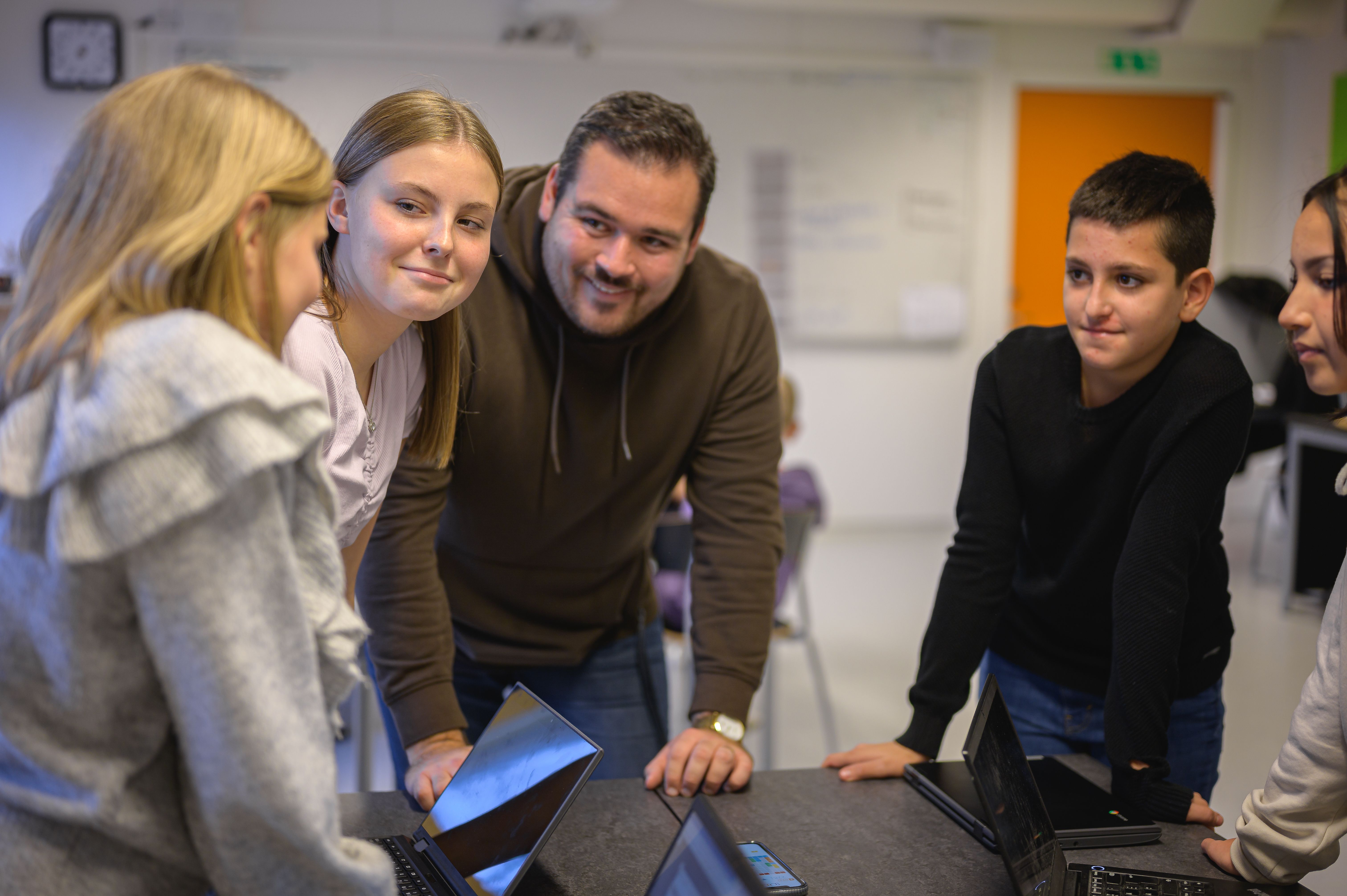 Lärare står omgiven av 4 elever vid datorer kring ett bord