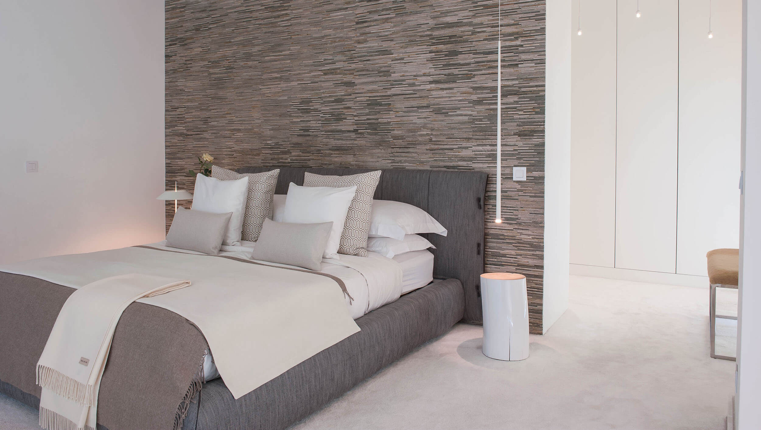 Grey upholstered bed, stacked stone feature wall and white cylindrical bedside table — luxury master suite design