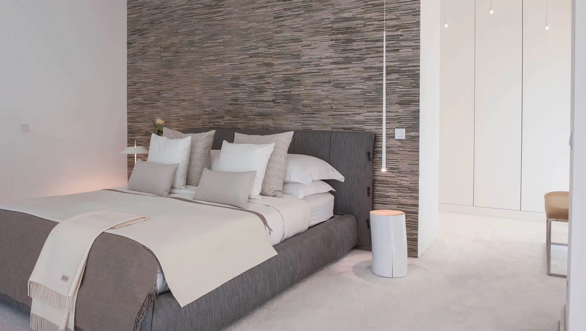 Grey upholstered bed, stacked stone feature wall and white cylindrical bedside table — luxury master suite design