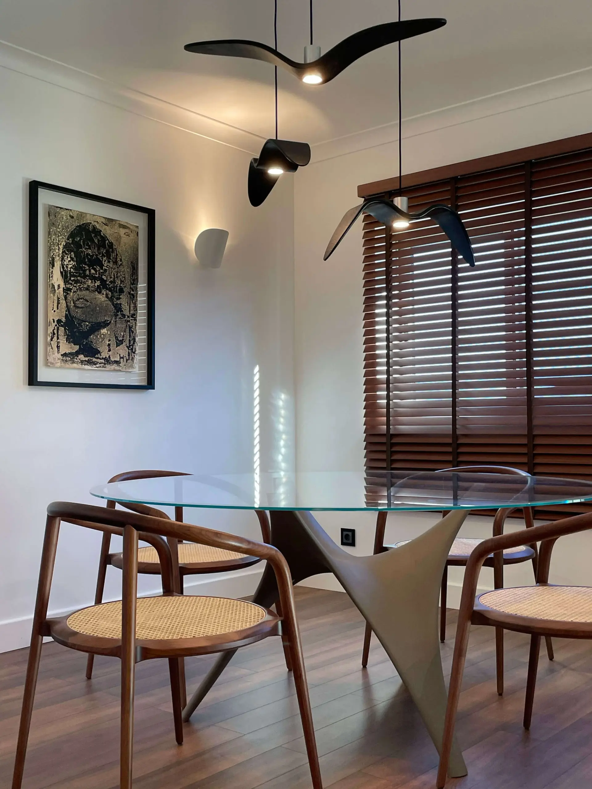 Round glass table on a sculptural curved base, cane-back wood armchairs and three black bird-form pendants — luxury dining room