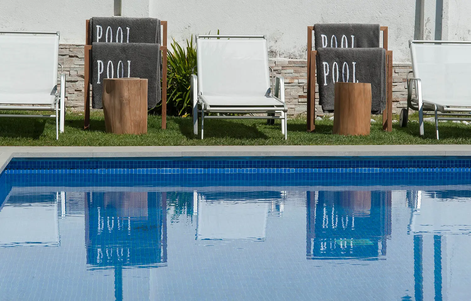 Detail of a blue pool with white loungers, personalized towels, and natural wood tables.