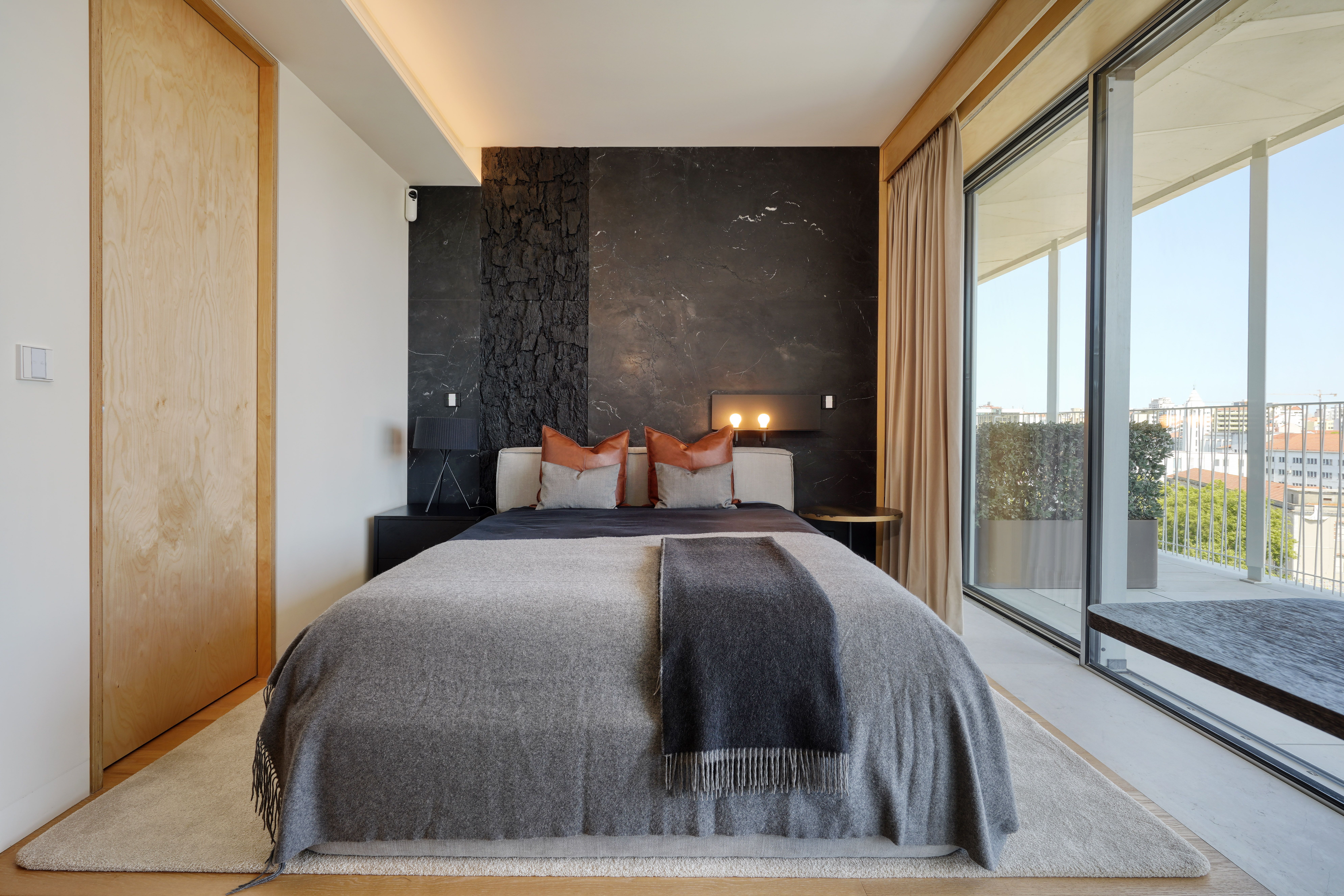 Suite com parede em pedra preta, cama em tons cinzentos e portas de madeira, num projeto do Atelier Renata Santos Machado em Lisboa.
